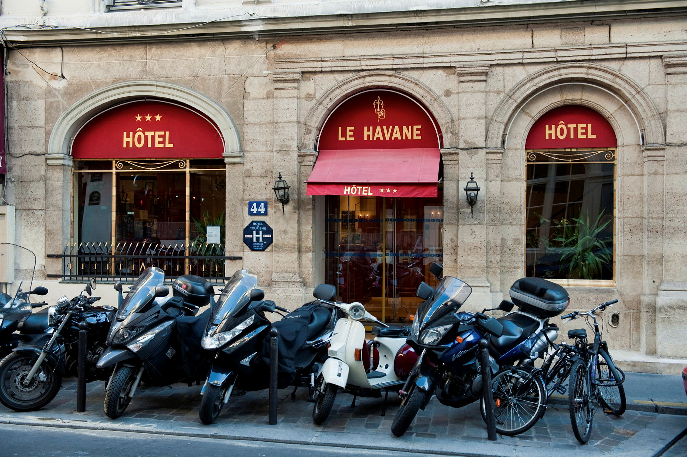 Bild som visar entrén till ett hotell med namnet Le Havane, tre stjärnor visas på markisen. Framför byggnaden står mopeder och cyklar parkerade. (AI-genererad text)