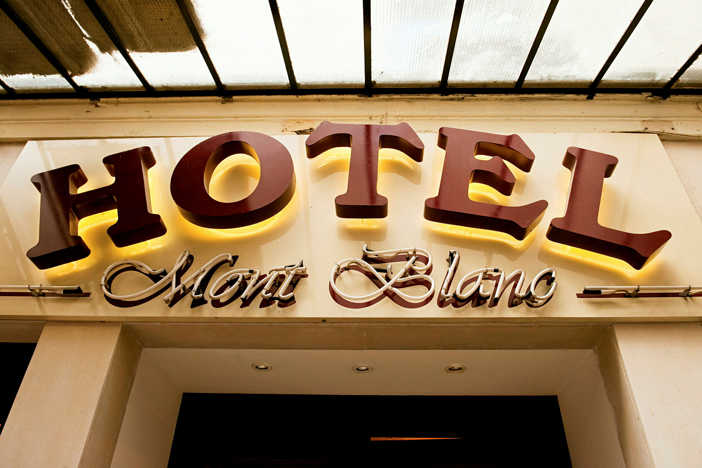 Ett hotellskylt med texten "HOTEL Mont Blanc", där "HOTEL" är i stora, belysta bokstäver och "Mont Blanc" är i en kursiv, dekorerad stil. (AI-genererad text)