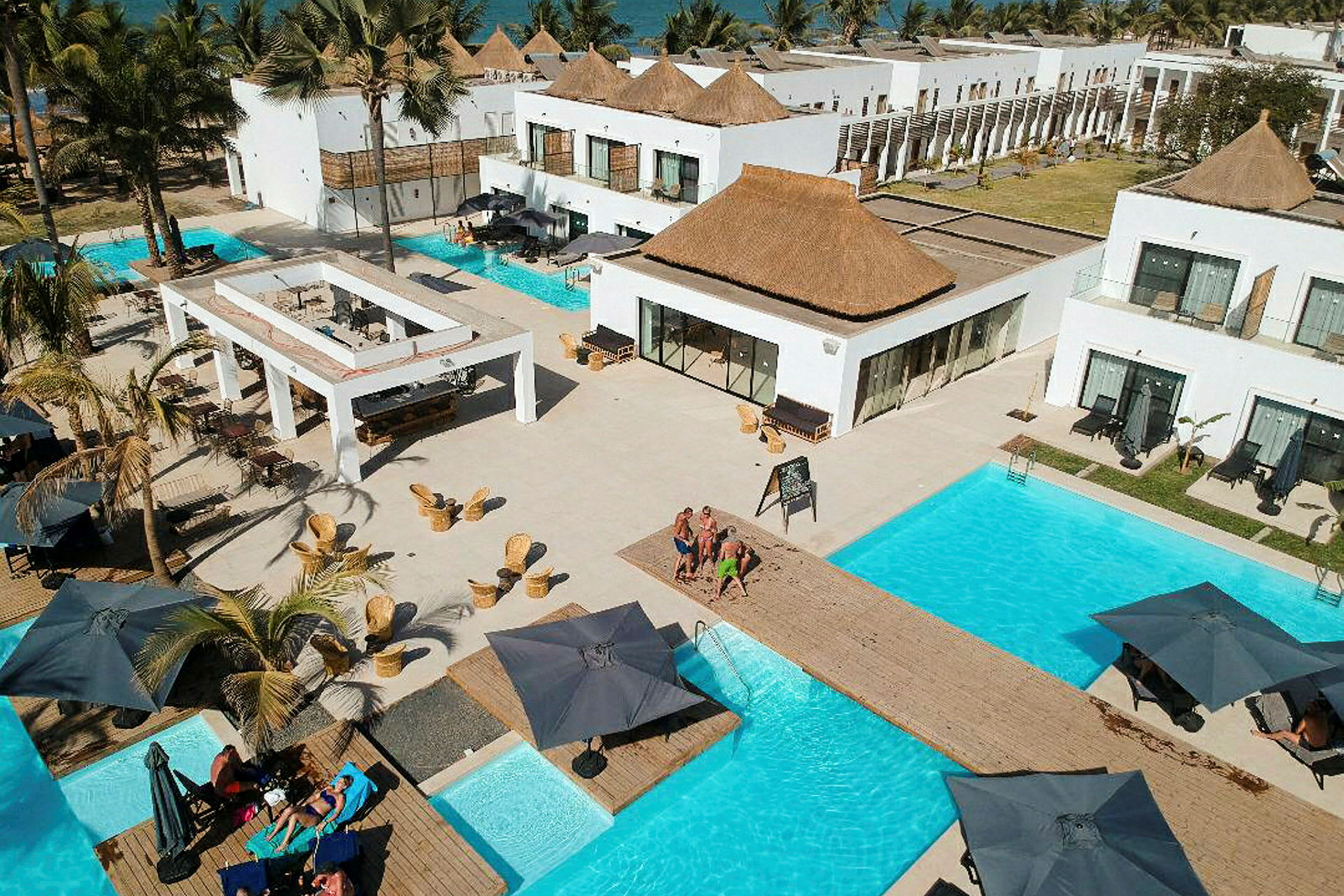 Vy över hotellanläggning med pooler, parasoller och vita byggnader med halmtak. (AI-genererad text)