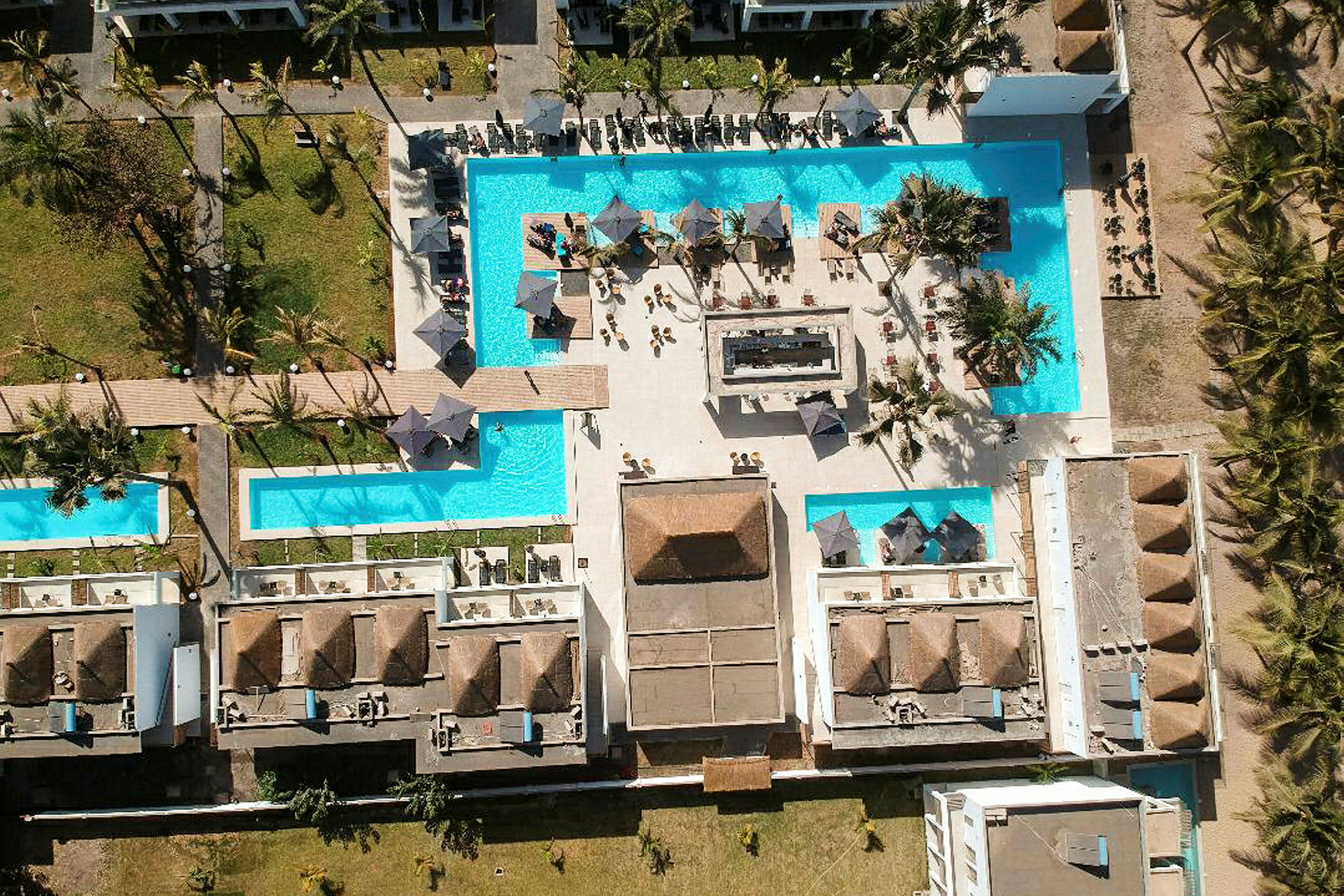 Flygfoto över en resort med swimmingpooler, solstolar och parasoller omgivna av byggnader och grönområden. (AI-genererad text)