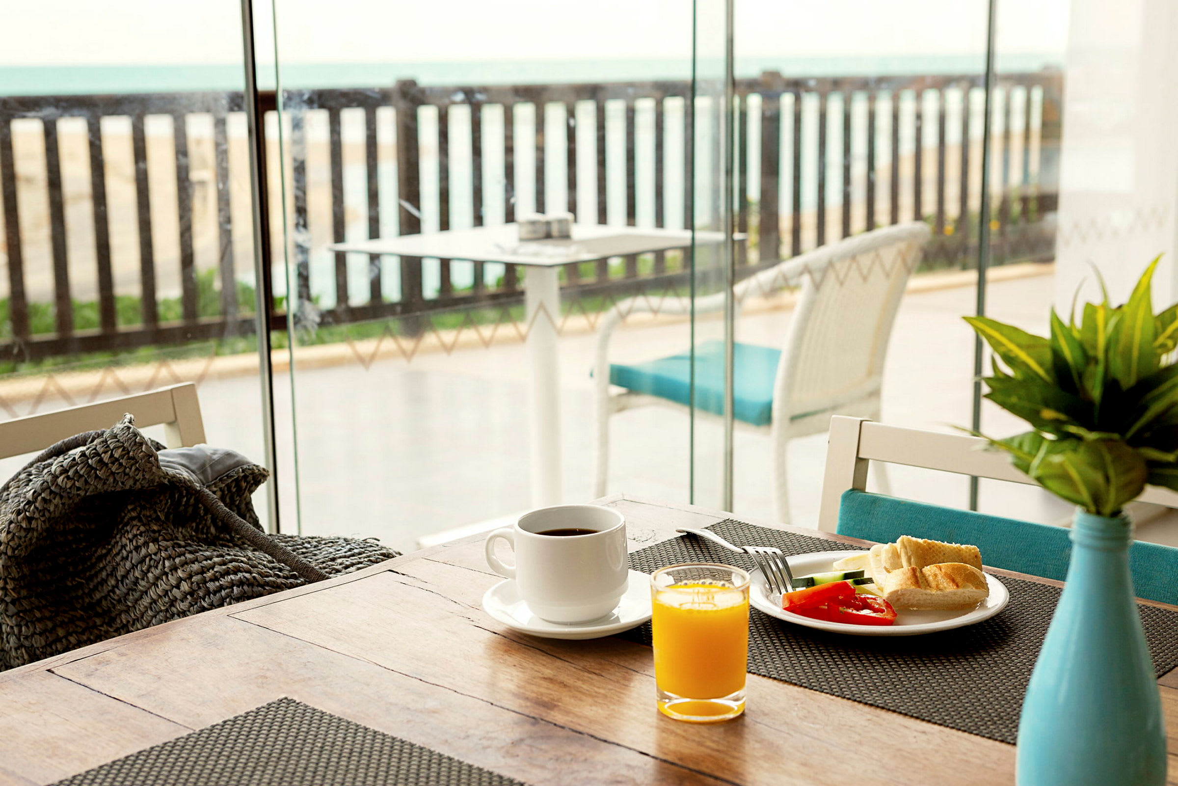 Frukostbord med kaffe, juice och grönsaker nära ett glasfönster med utsikt över en terrass med havet i bakgrunden. (AI-genererad text)