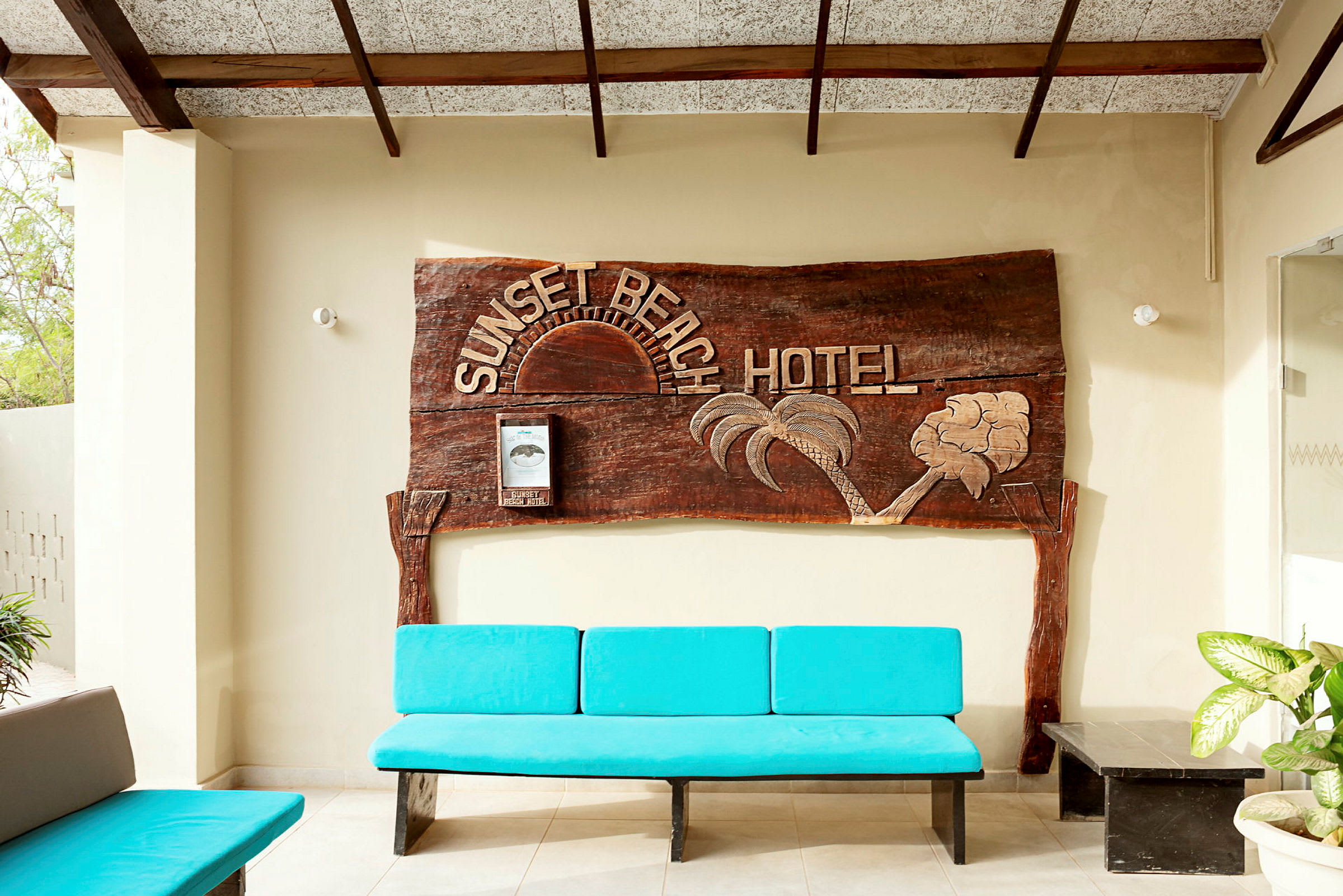 En träskylt med texten "Sunset Beach Hotel" pryder väggen. Framför finns en blå bänk och några växter. (AI-genererad text)