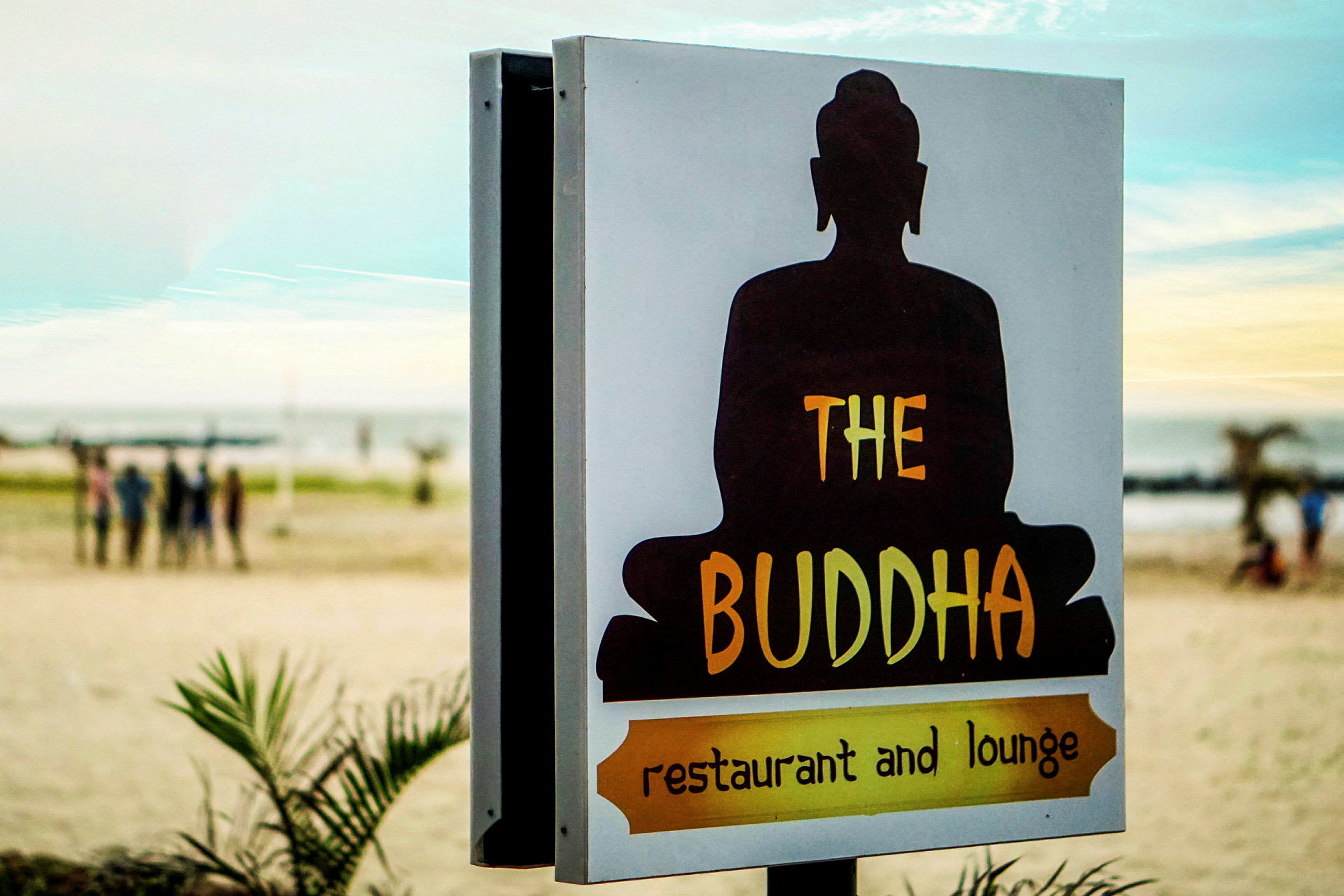 En skylt med texten 'The Buddha restaurant and lounge' med en silhuett av en sittande Buddha framför en strandbakgrund. (AI-genererad text)