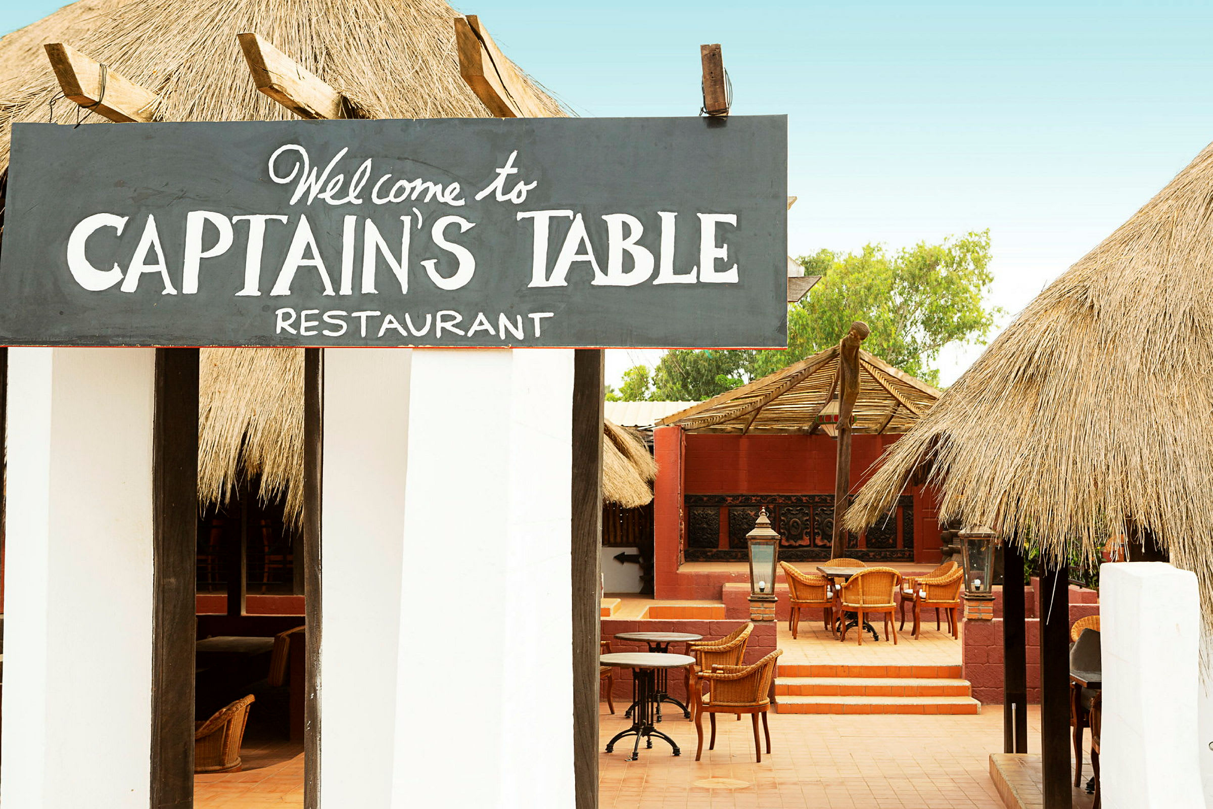 Ett restaurangområde med skylttexten "Welcome to Captain's Table Restaurant", stolar och bord synliga utomhus under tak. (AI-genererad text)