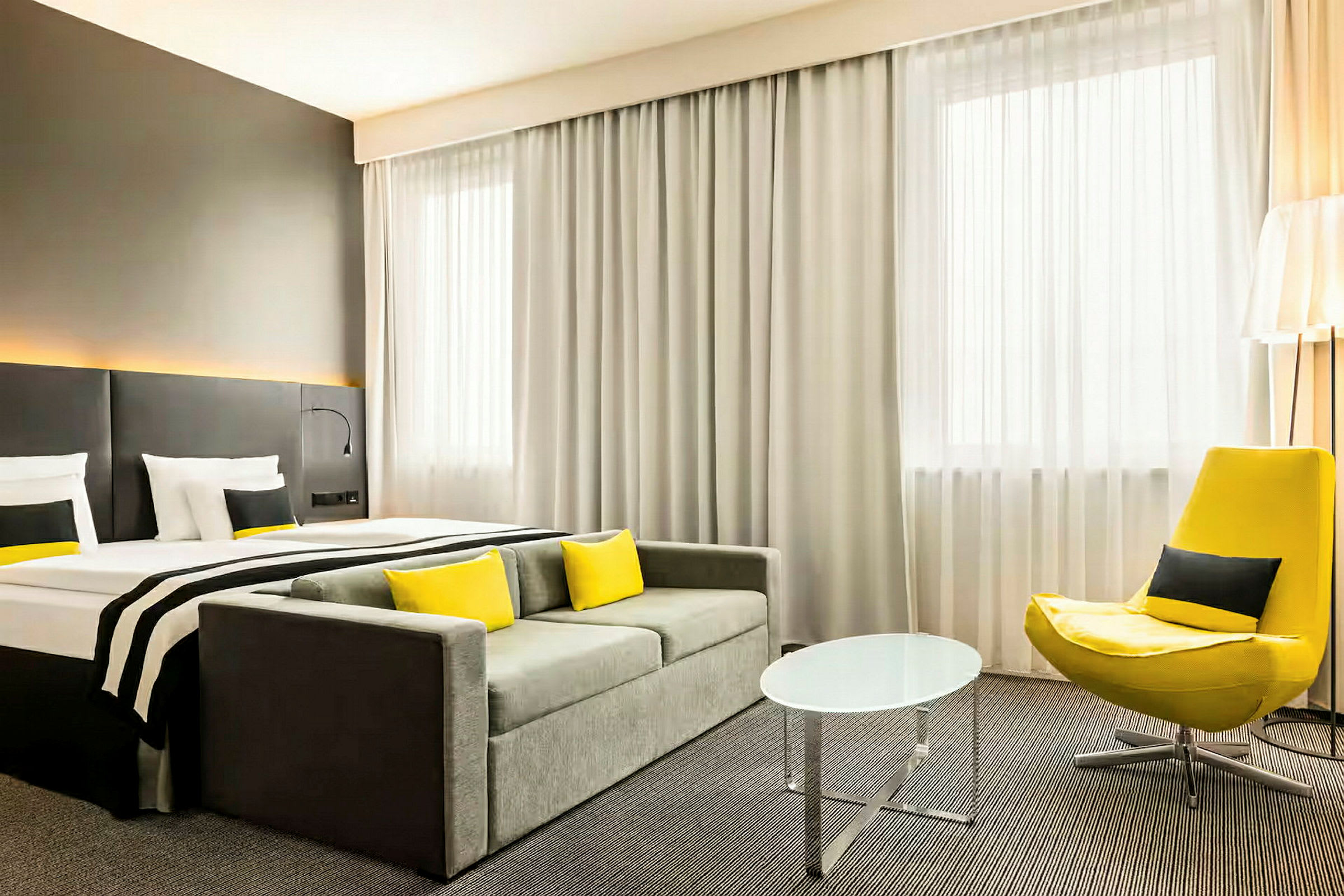 Ett hotellrum med en säng, soffa, gul fåtölj och ett litet runt bord. (AI-genererad text)