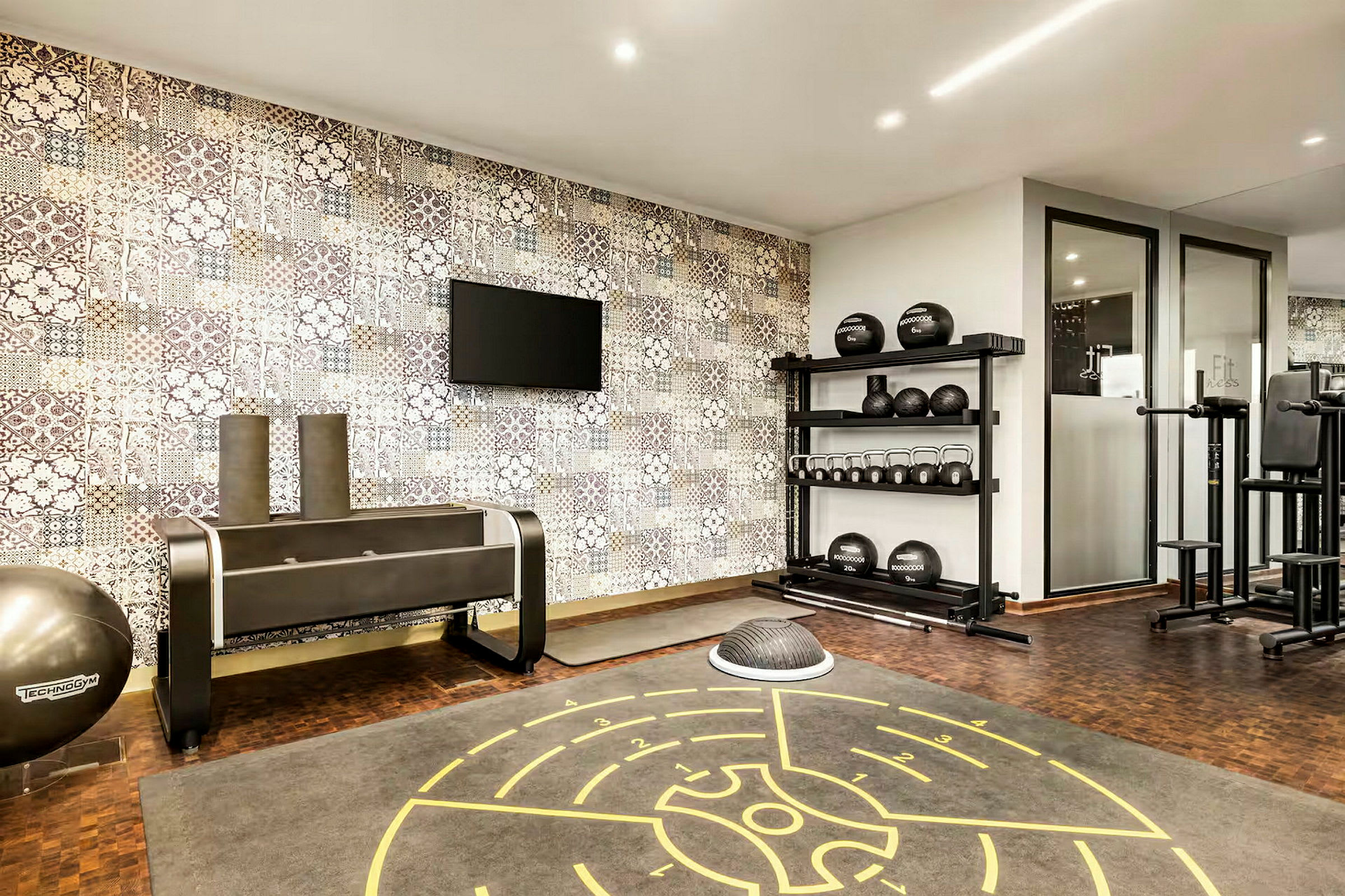 Ett gym med träningsutrustning så som kettlebells, medicinbollar och en träningsmatta på golvet. En tv är monterad på väggen. (AI-genererad text)