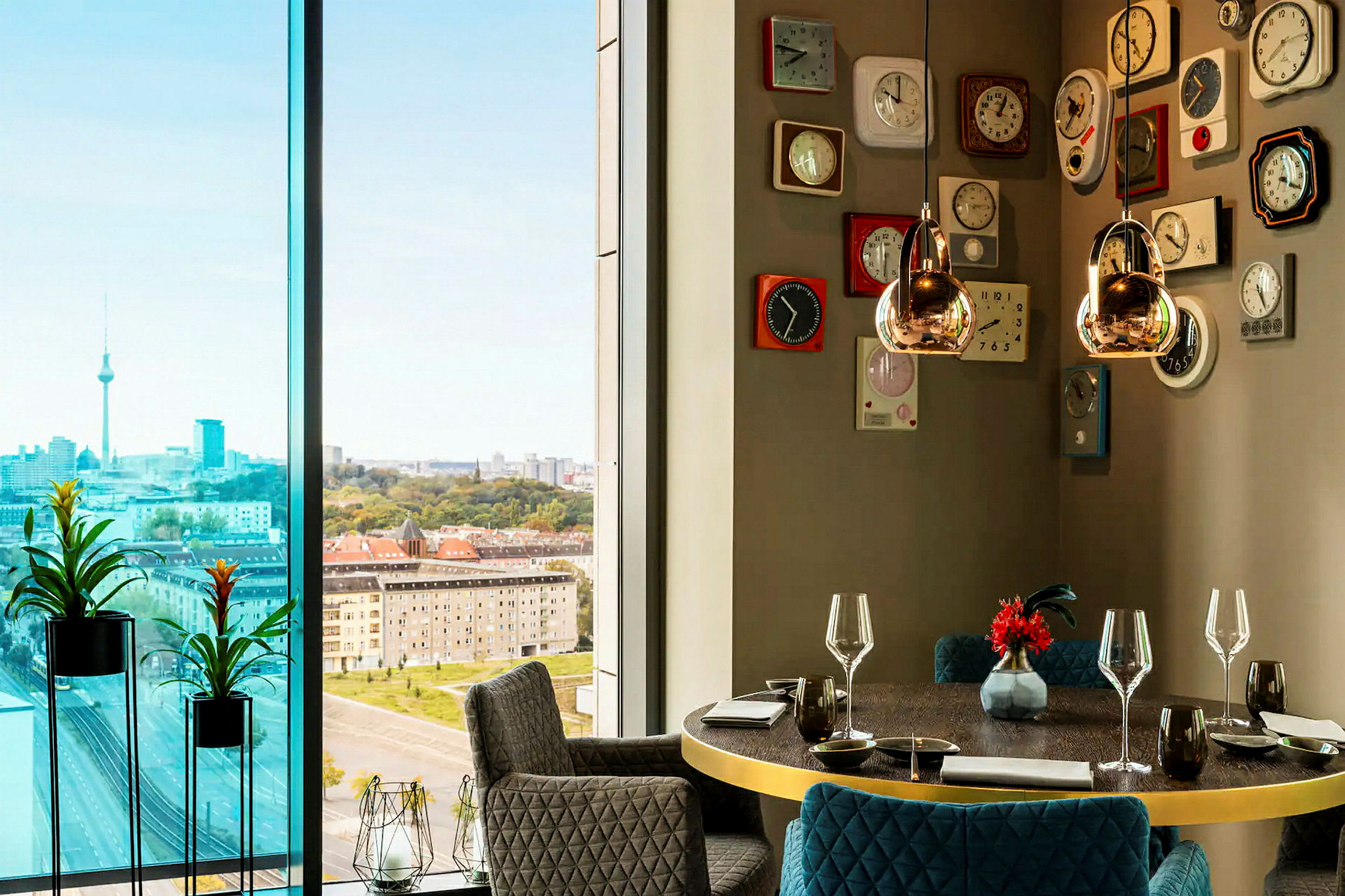 Ett modernt rum med stora fönster som visar en urban utsikt med torn, inramat av höga byggnader och grönska. Interiören inkluderar en rund matbord med glas och porslin samt en vägg dekorerad med retro-inspirerade klockor. (AI-genererad text)