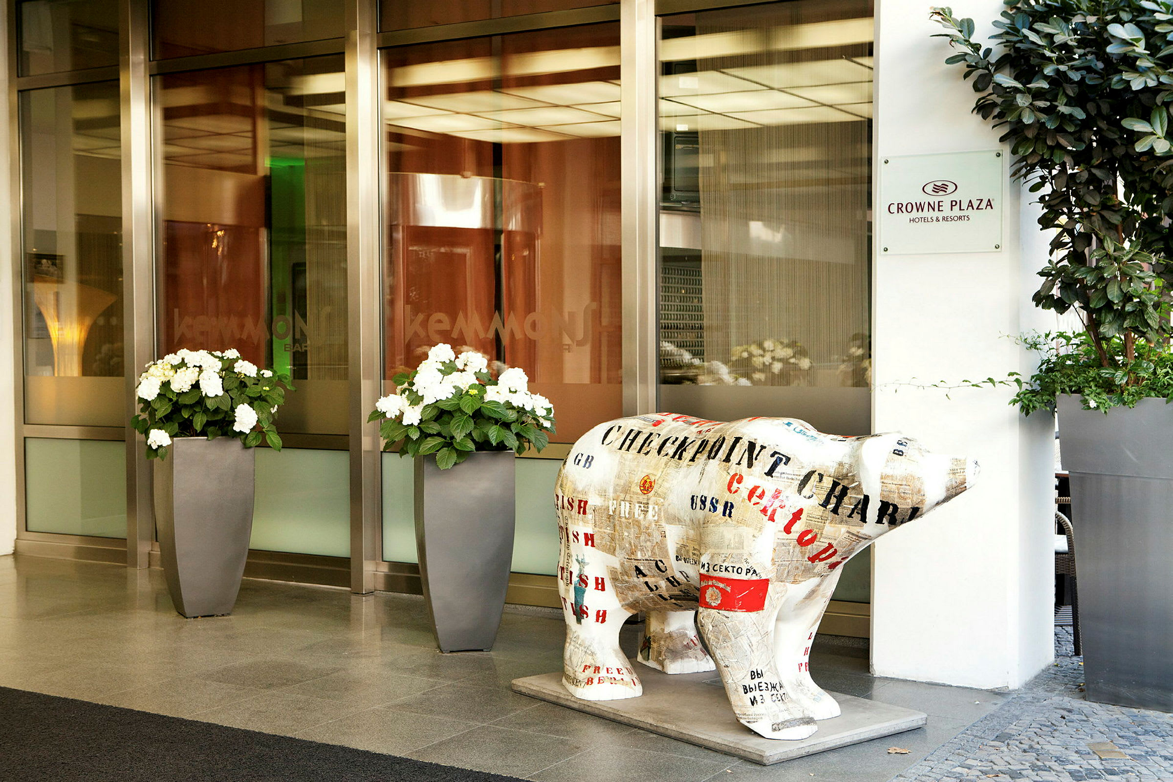 Utsidan av hotellet Crowne Plaza med blomsterkrukor och en björnstaty dekorerad med text och motiv. (AI-genererad text)
