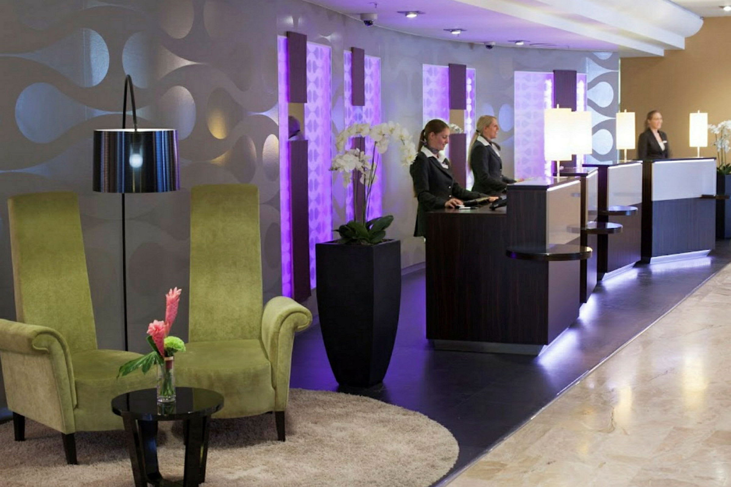 En hotellobby med två gröna stolar, ett litet bord med en blomma, och receptioner med belysning. (AI-genererad text)
