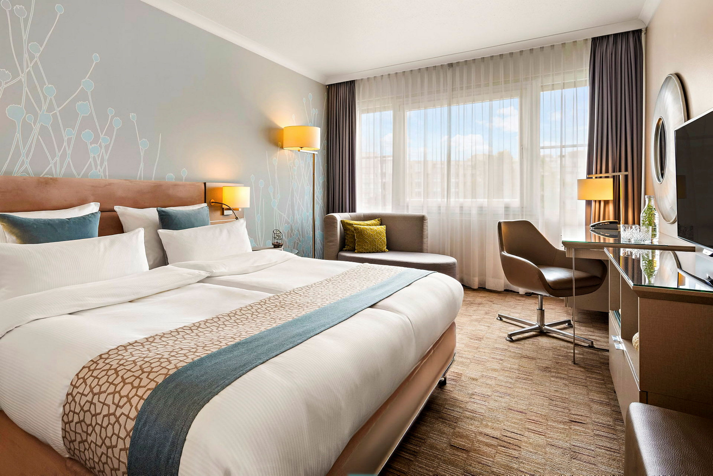 Ett modernt hotellrum med en stor dubbelsäng, skrivbord, stol och en sittgrupp vid ett fönster. (AI-genererad text)