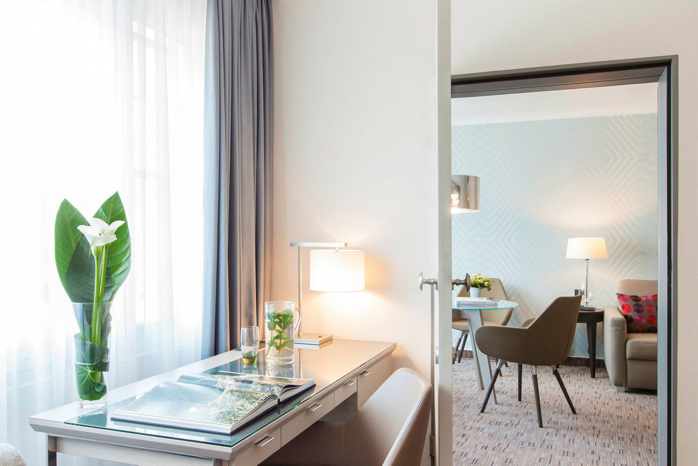 Ett hotellrum med ett skrivbord, lampa och en blomvas, samt en sittgrupp i bakgrunden. (AI-genererad text)