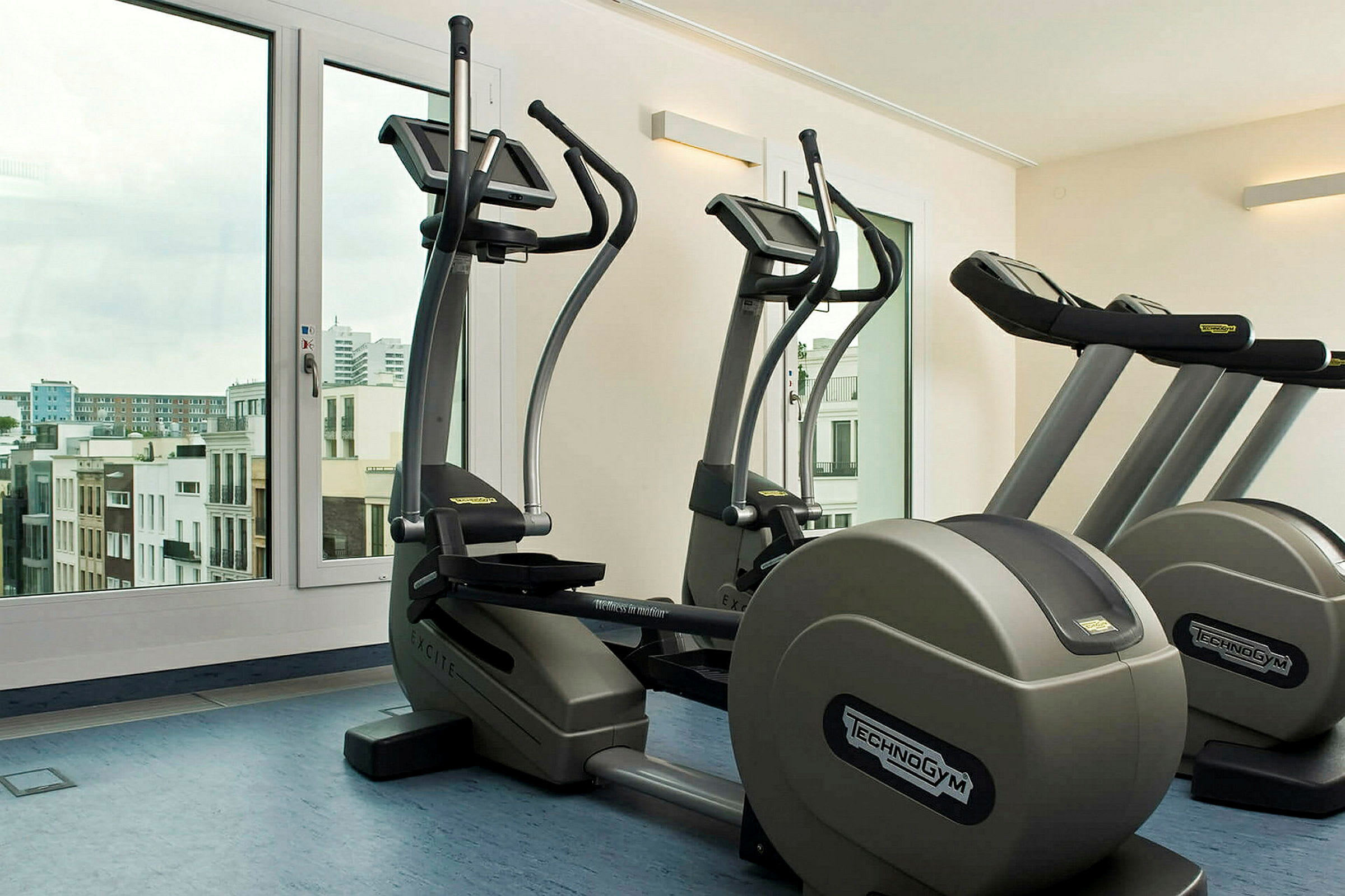 Ett gym med träningscyklar och löpband framför stora fönster med utsikt över en stad. (AI-genererad text)