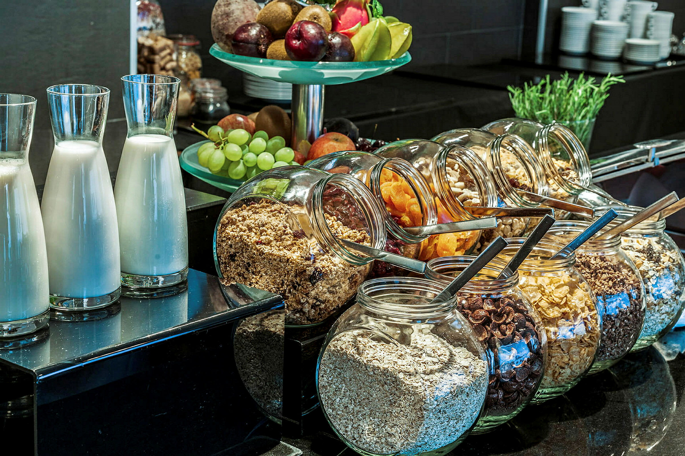 En frukostbuffé med glasflaskor med mjölk, glasburkar med olika typer av flingor och müsli, samt en skål med färsk frukt. (AI-genererad text)