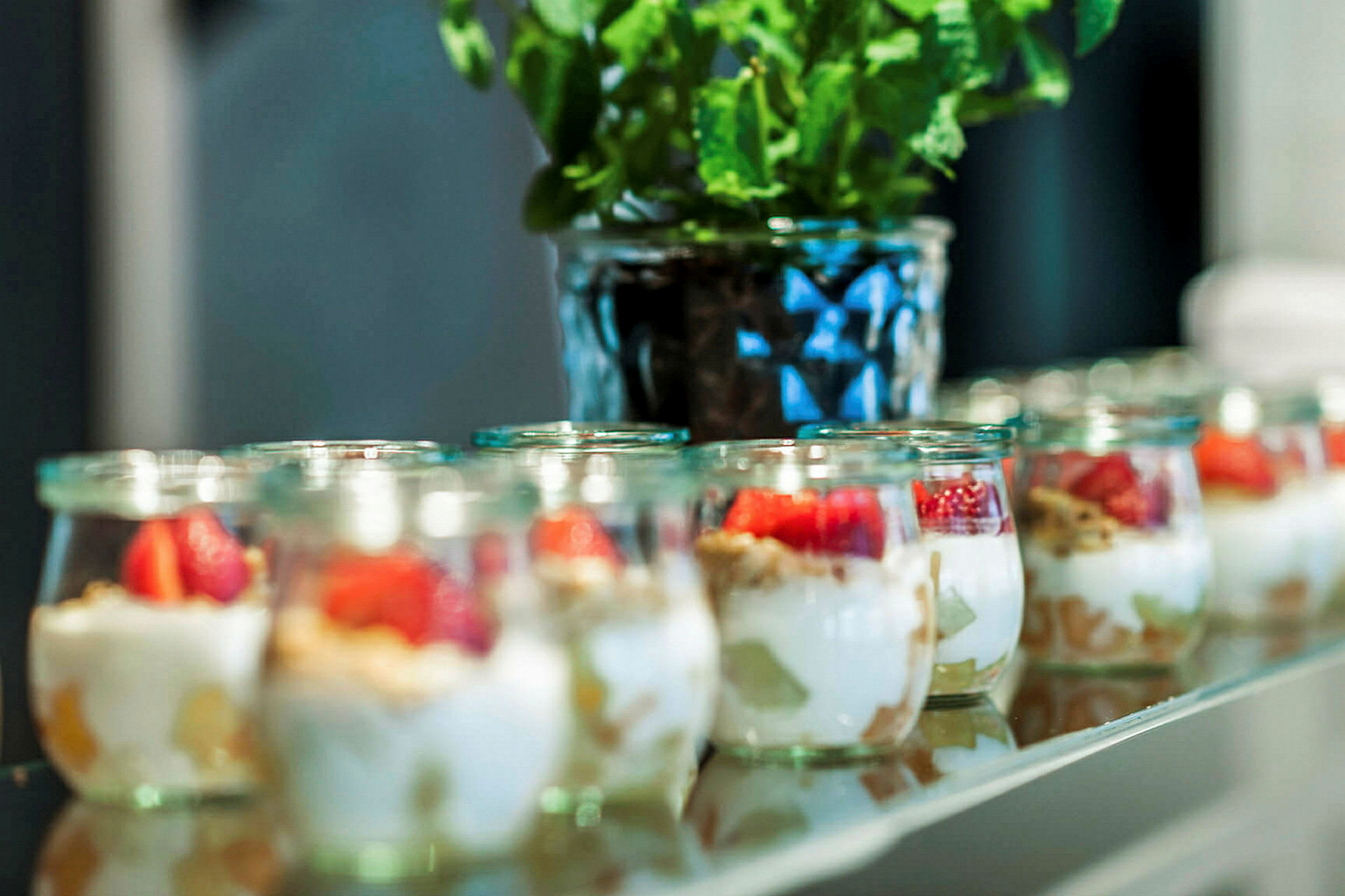 Små glasburkar med yoghurt, frukt och granola står uppställda på en rad. I bakgrunden syns en krukväxt. (AI-genererad text)