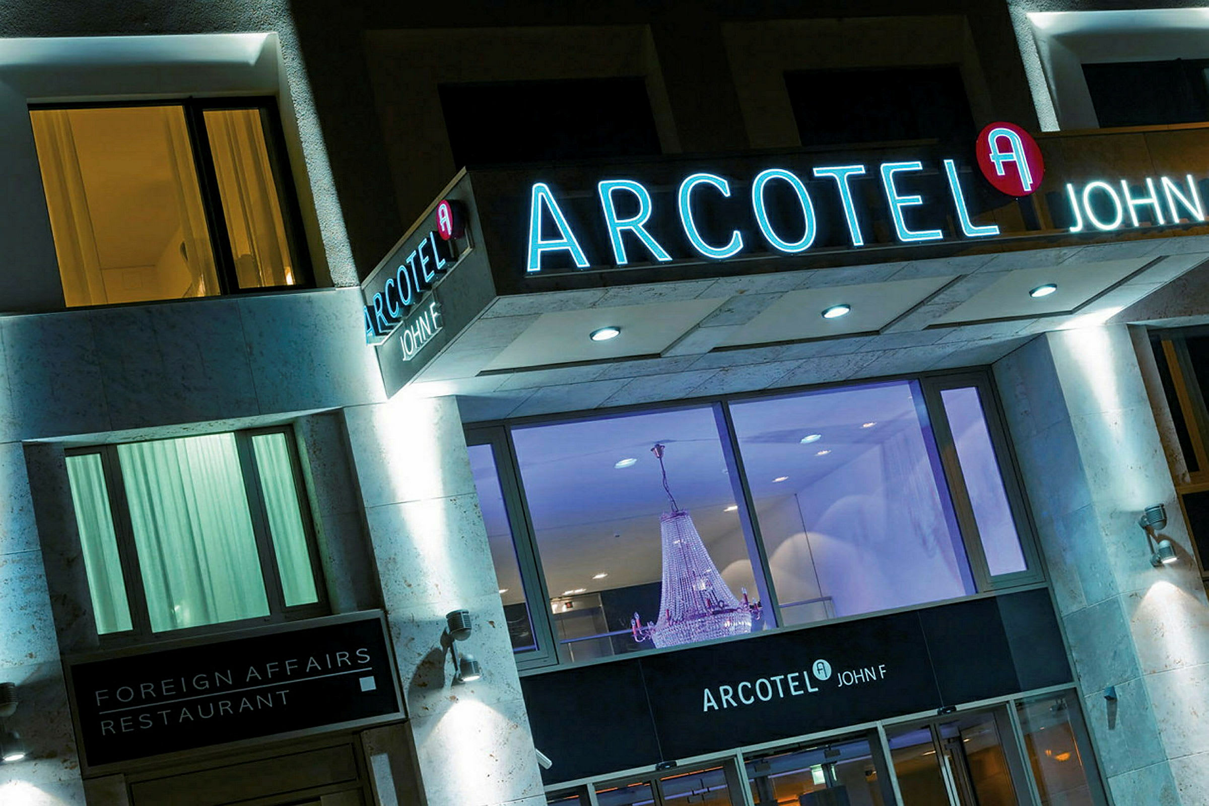 Bild av en hotellfasad med ljusskyltar som visar namnet Arcotel John F, samt en restaurang kallad Foreign Affairs. En stor kristallkrona syns genom de stora fönstren. (AI-genererad text)