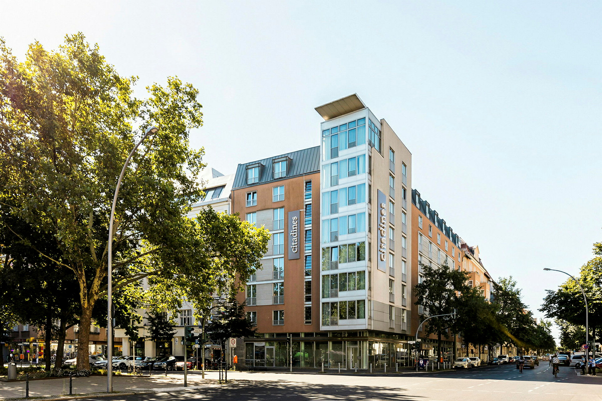 Ett modernt hotell med glasfasad omgiven av träd och stadsmiljö. (AI-genererad text)