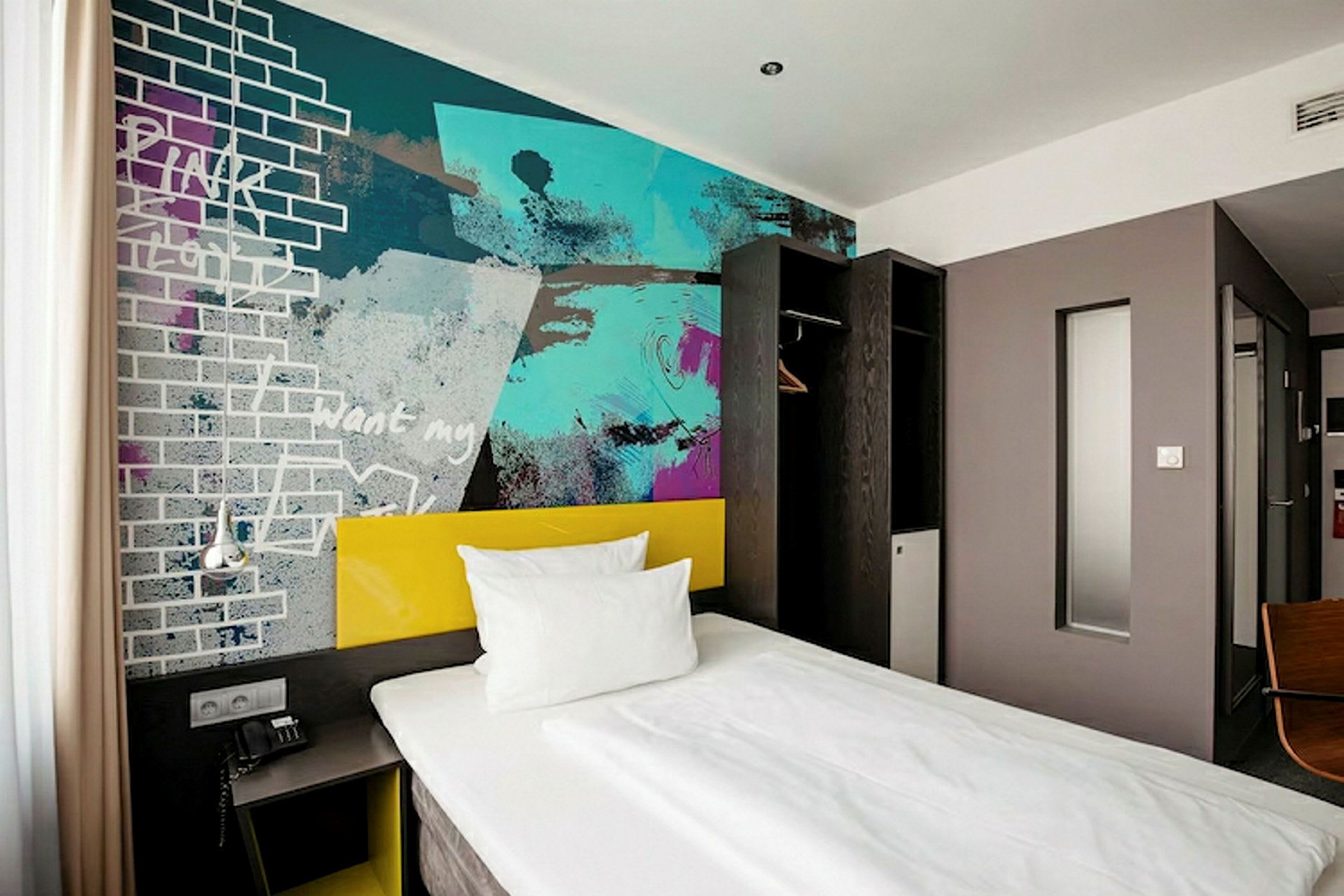 Ett hotellrum med en enkel säng, en färgglad väggmålning och minimalistisk inredning. (AI-genererad text)