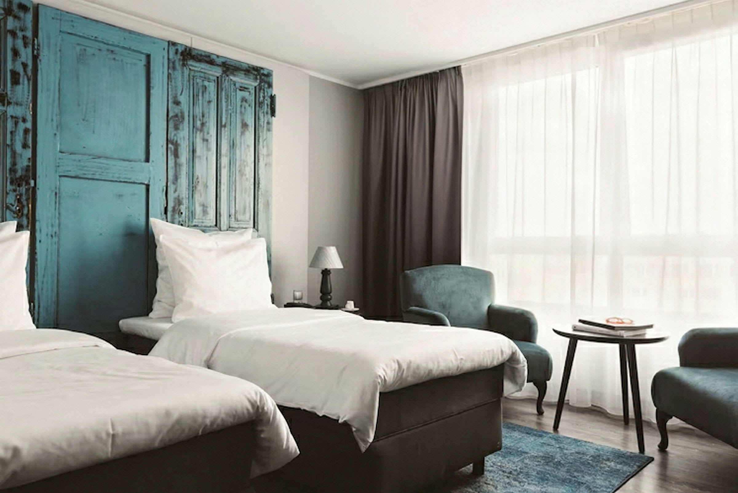 Ett hotellrum med två enkelsängar, blå detaljer på väggen, ett bord och fåtöljer, samt ett stort fönster med vit gardin. (AI-genererad text)