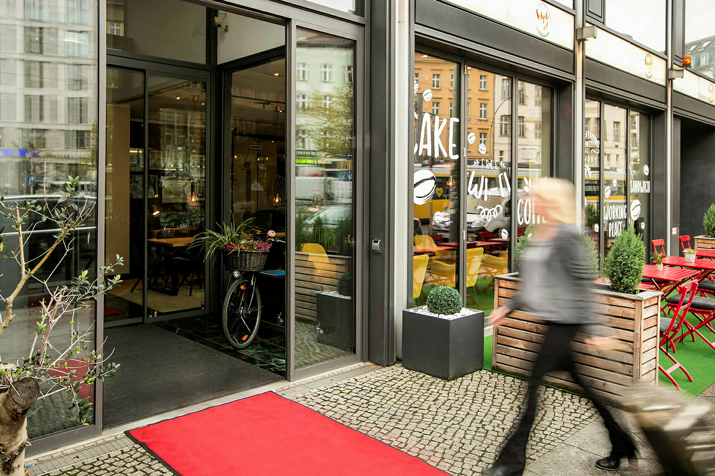 Entré till ett kafé med glasfasad, röd matta på marken, och en person som passerar utanför. (AI-genererad text)
