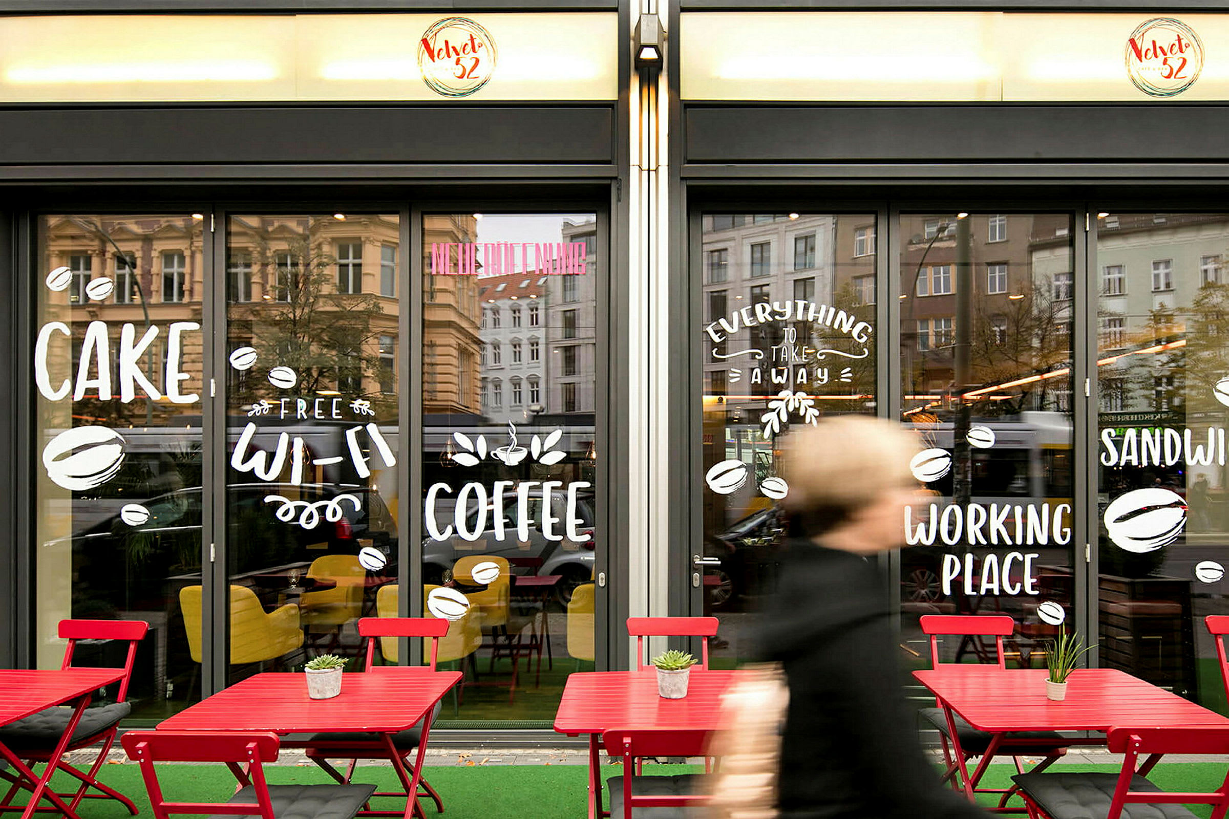 Framsidan av ett kafé med stora glasfönster, dekorerade med text och symboler. Utanför står röda bord och stolar. (AI-genererad text)