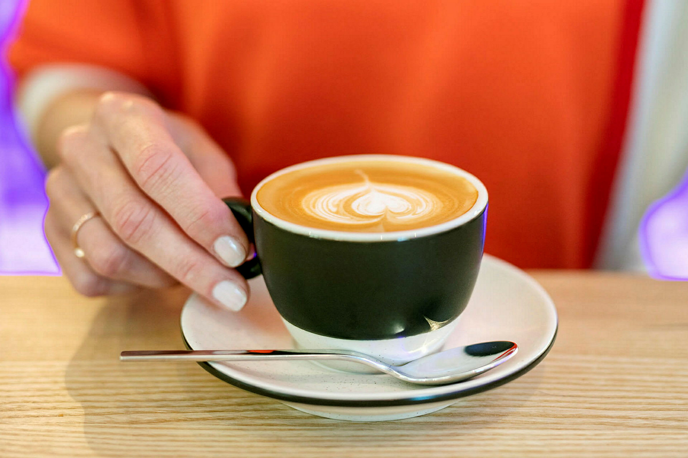 En hand håller i ett svart kaffekoppsöra med ett fat och sked på ett träbord. Cappuccino med latte art på toppen. (AI-genererad text)