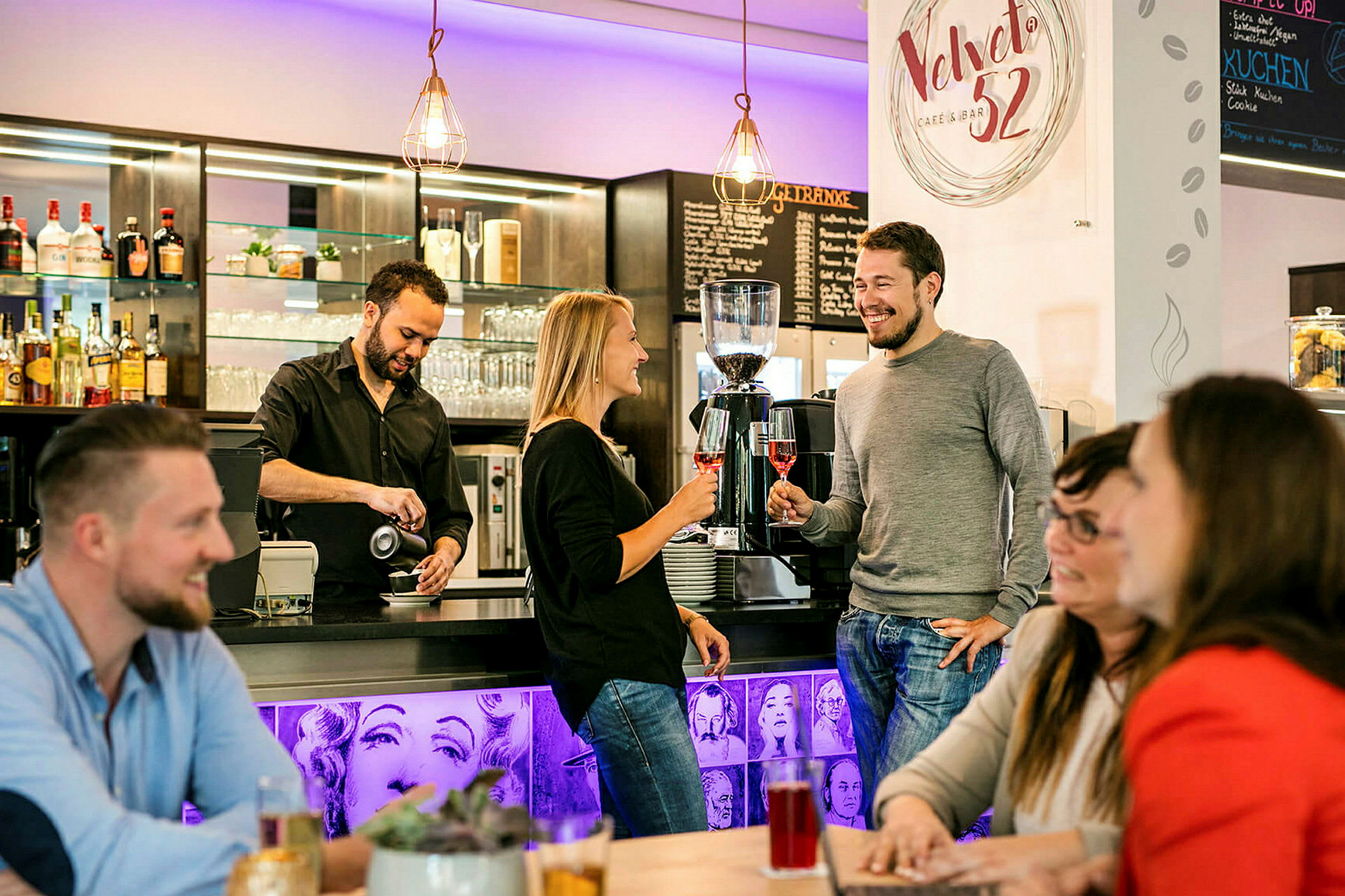 En barista häller kaffe vid en bardisk där människor står och sitter och njuter av sina drycker. (AI-genererad text)