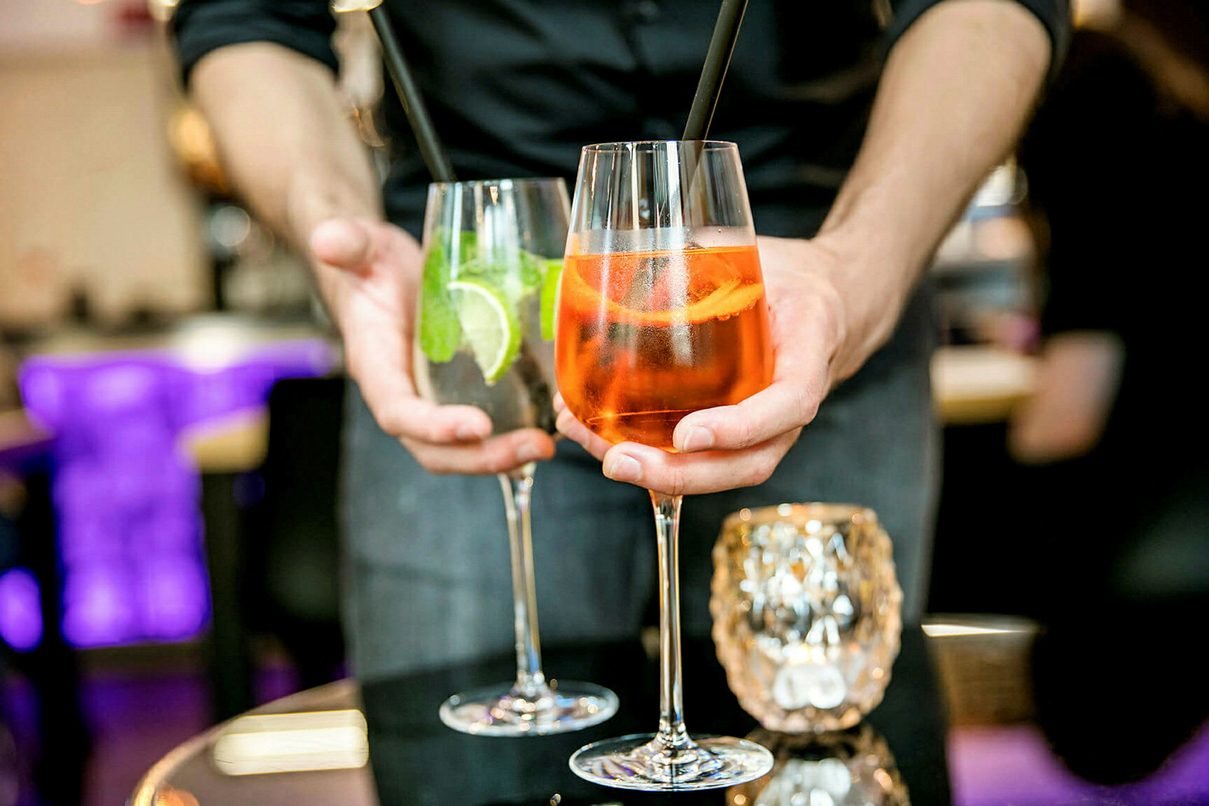 Två drinkar hålls fram av en person, ett glas innehåller en orange cocktail och det andra en klar cocktail med lime och mynta. (AI-genererad text)