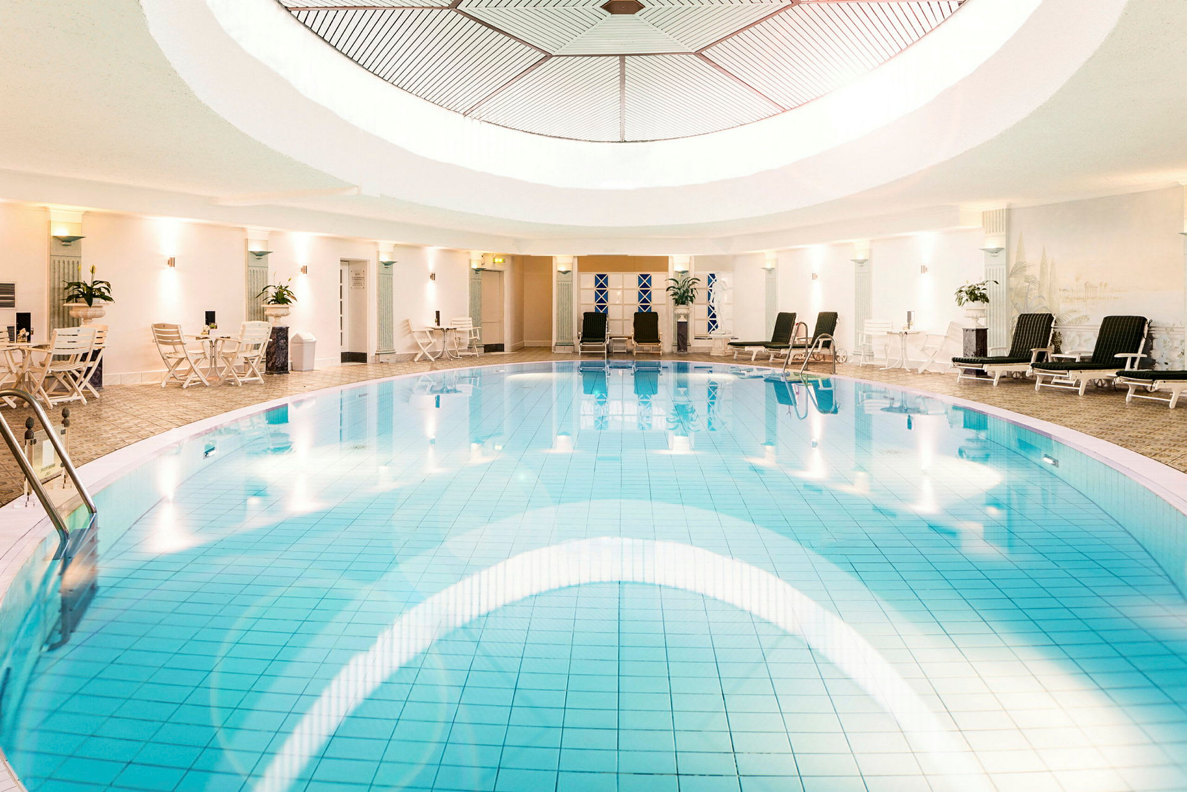 En inomhuspool med klarblått vatten, omgiven av solstolar och små bord i en ljus och elegant miljö. (AI-genererad text)