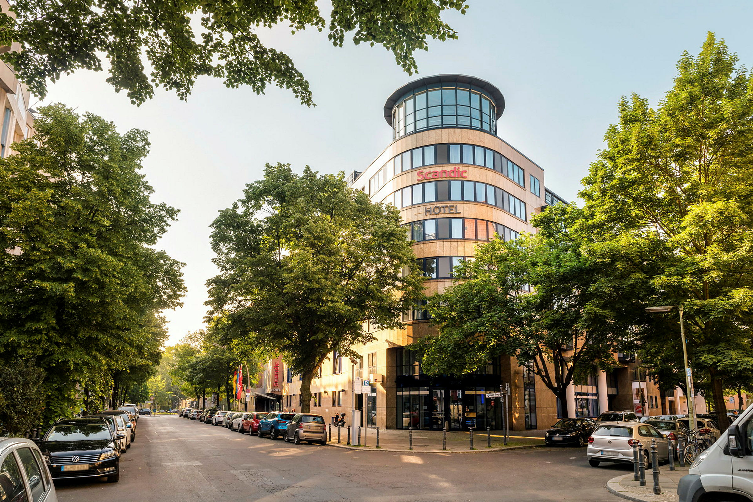 En stadsbild med fasaden av ett Scandic hotell omgiven av träd och parkerade bilar. (AI-genererad text)