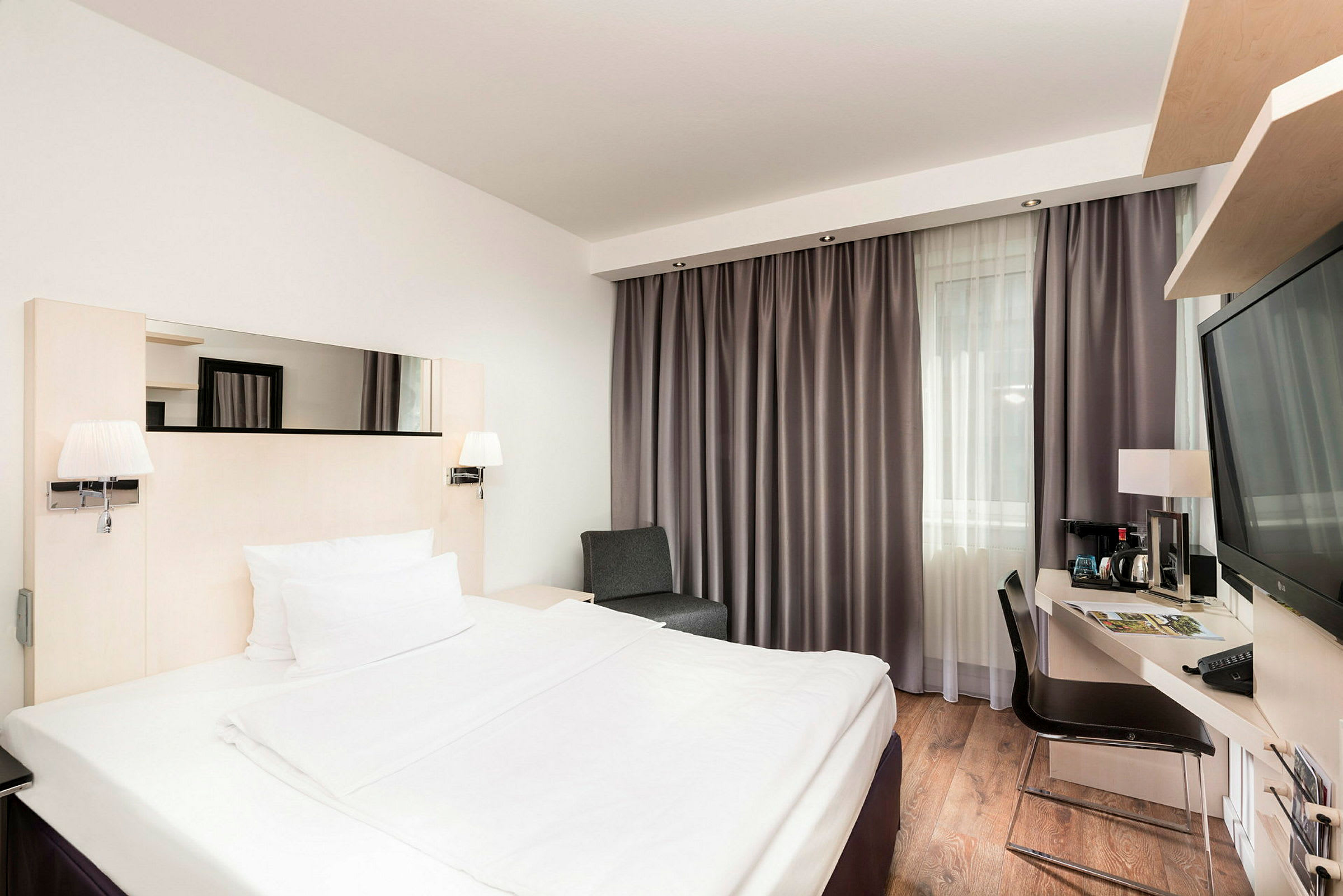 Ett hotellrum med en dubbelsäng, skrivbord och stol, samt ett stort fönster med gardiner. (AI-genererad text)