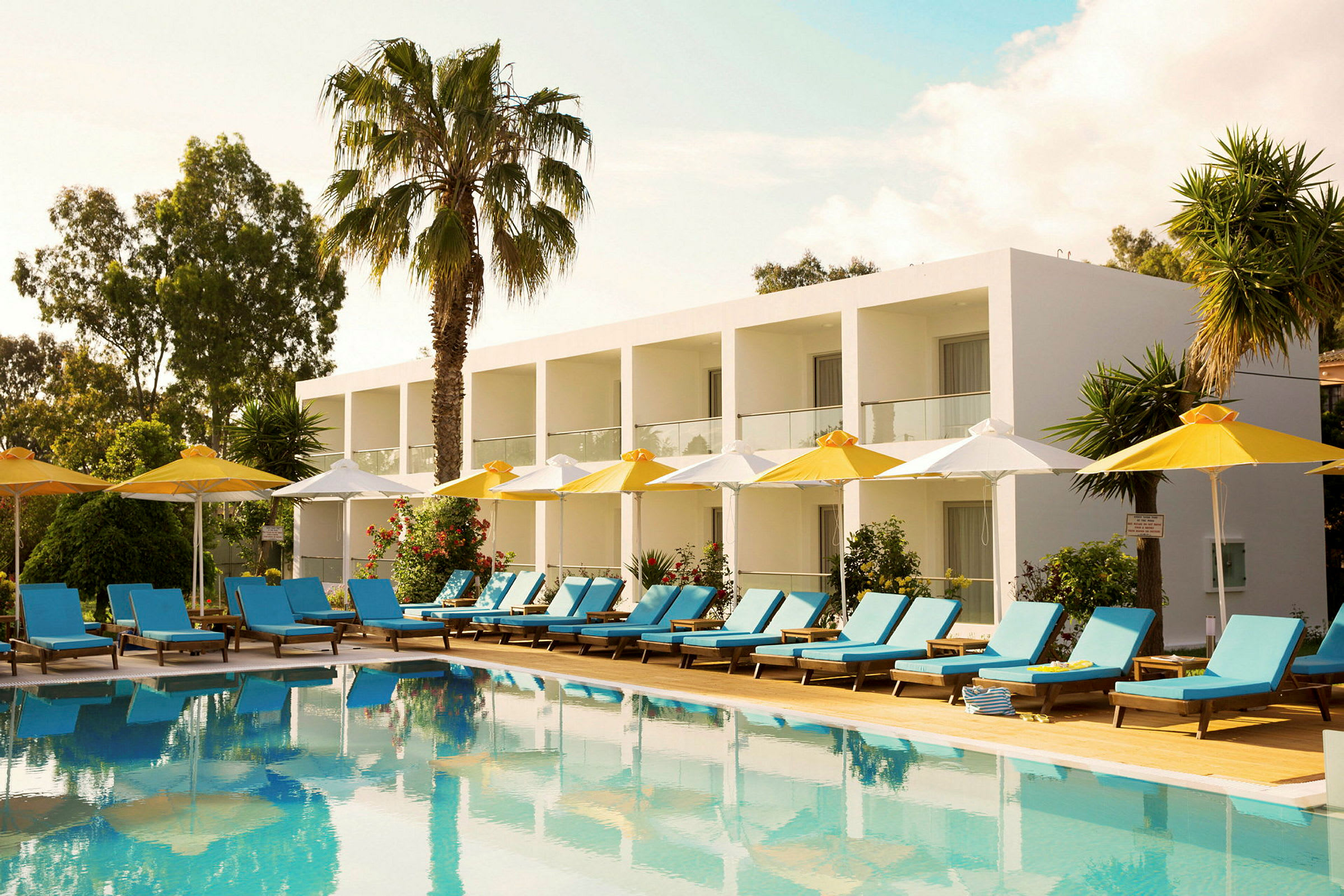 Ett hotell med vita byggnader och balkonger i en grön omgivning, framför en pool med solstolar och parasoller. (AI-genererad text)