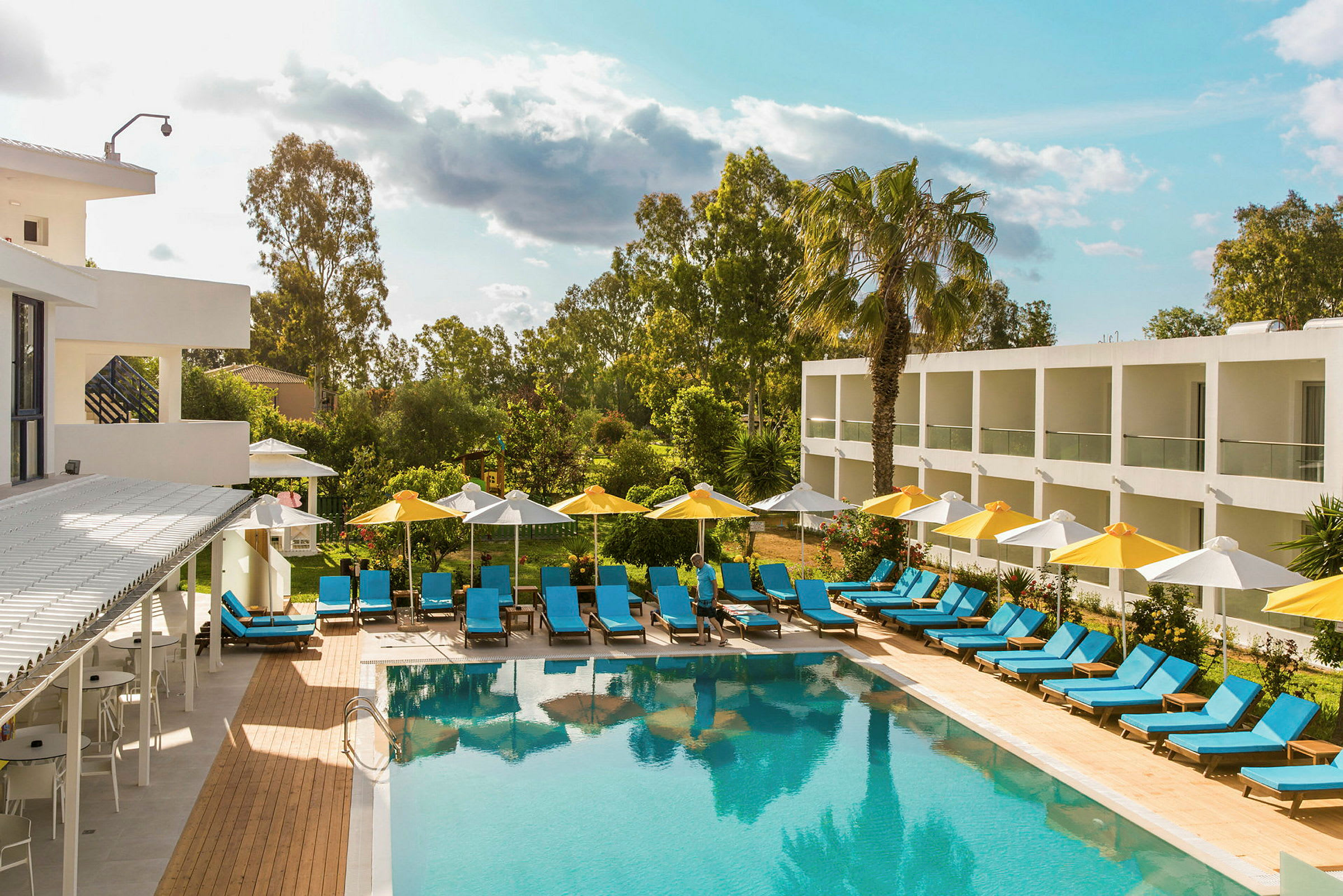 Ett hotellområde med en utomhuspool omgiven av solstolar och parasoller. Byggnader och grönska syns i bakgrunden. (AI-genererad text)