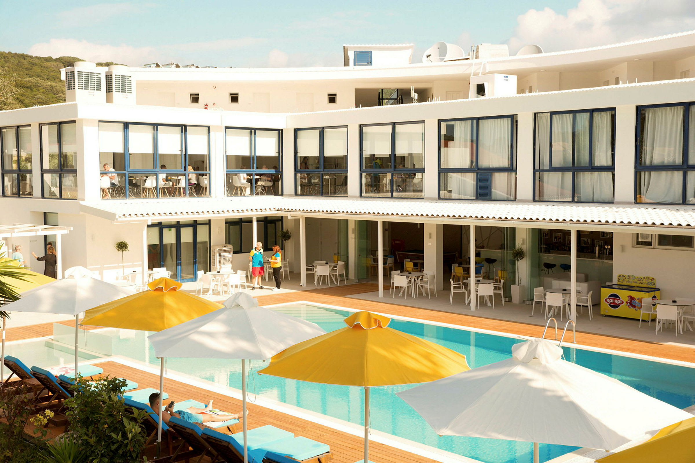 En bild av en hotellbyggnad med pool och solstolar, samt parasoller i gult och vitt. (AI-genererad text)