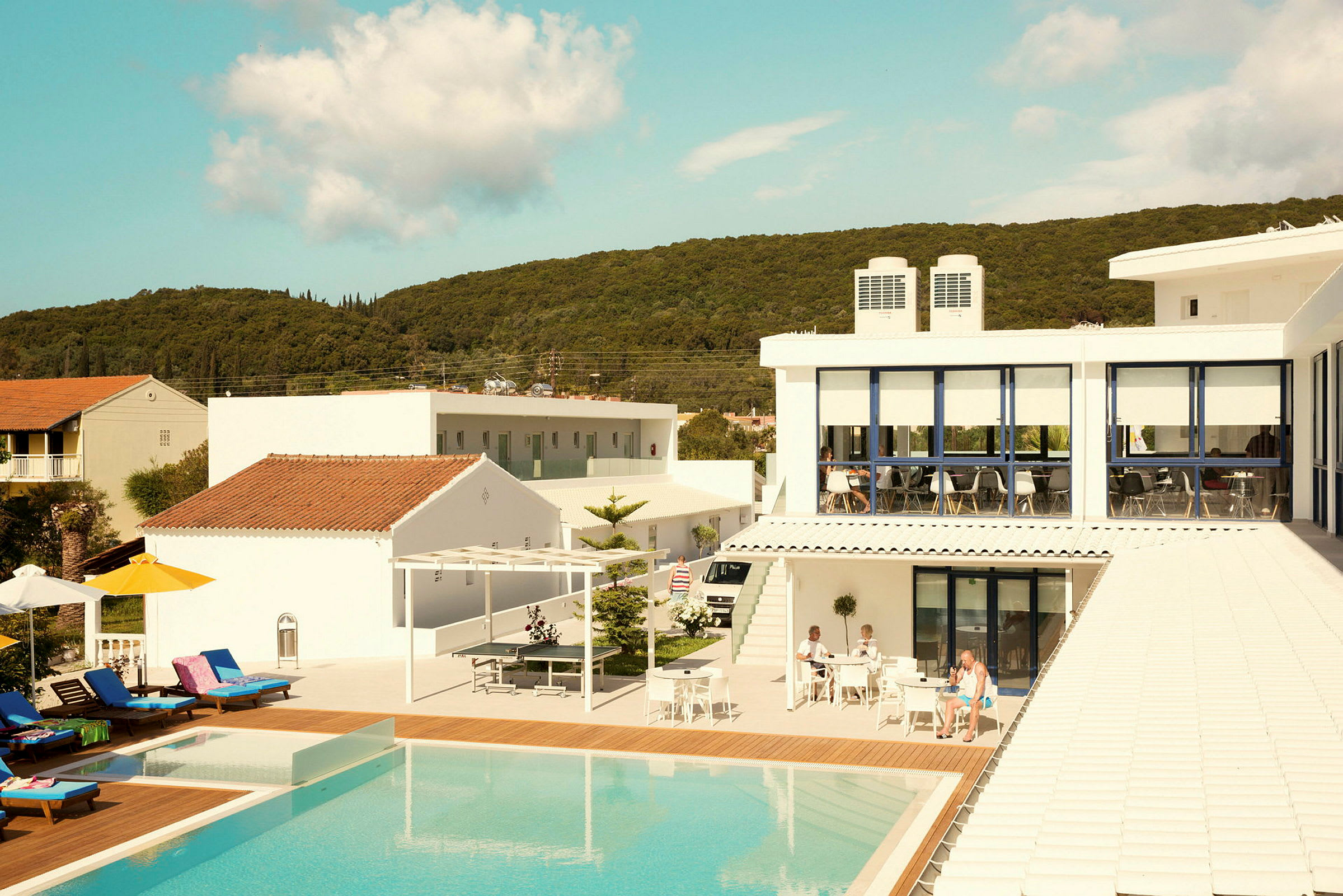 Ett hotell med en pool och solstolar, omgiven av byggnader och natur. (AI-genererad text)