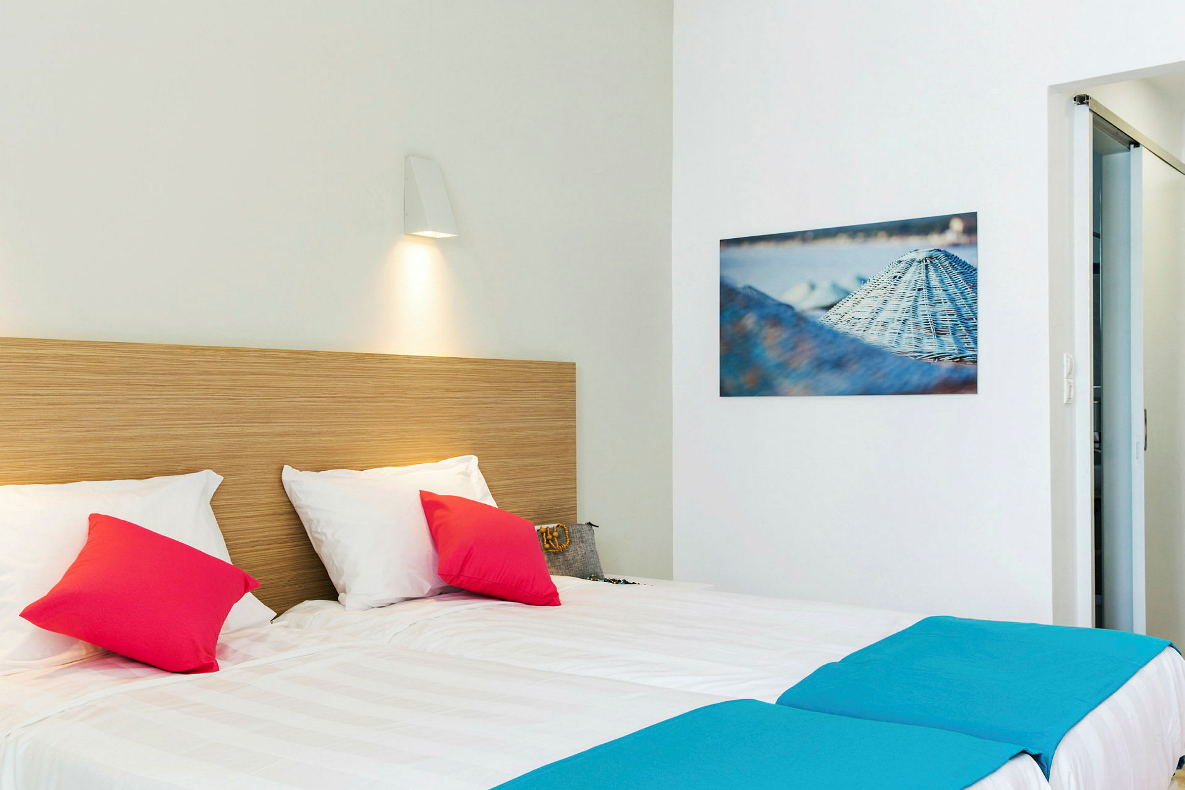 Hotellrum med dubbelsäng bäddad med vita lakan, dekorativa färgglada kuddar och en träpanel som sänggavel. På väggen hänger en tavla och en lampa är riktad ned mot sängen. (AI-genererad text)