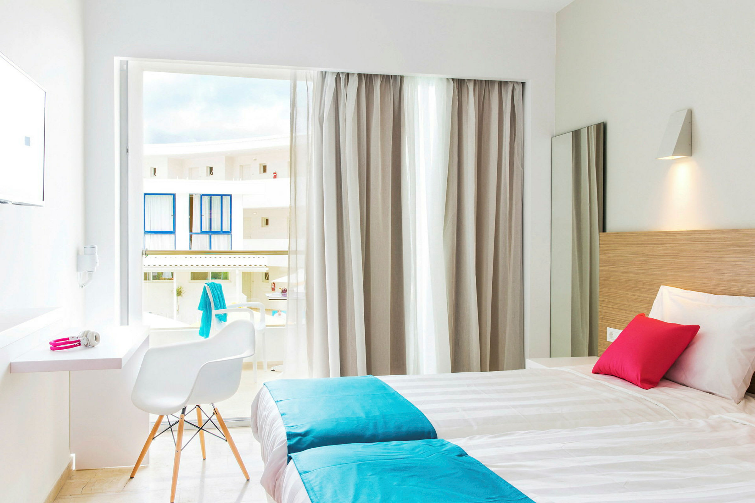Ett ljust hotellrum med två enkelsängar, vita och turkosa sängkläder, en rosa kudde. Fönstret har ljusa gardiner och öppnar mot balkonger. (AI-genererad text)