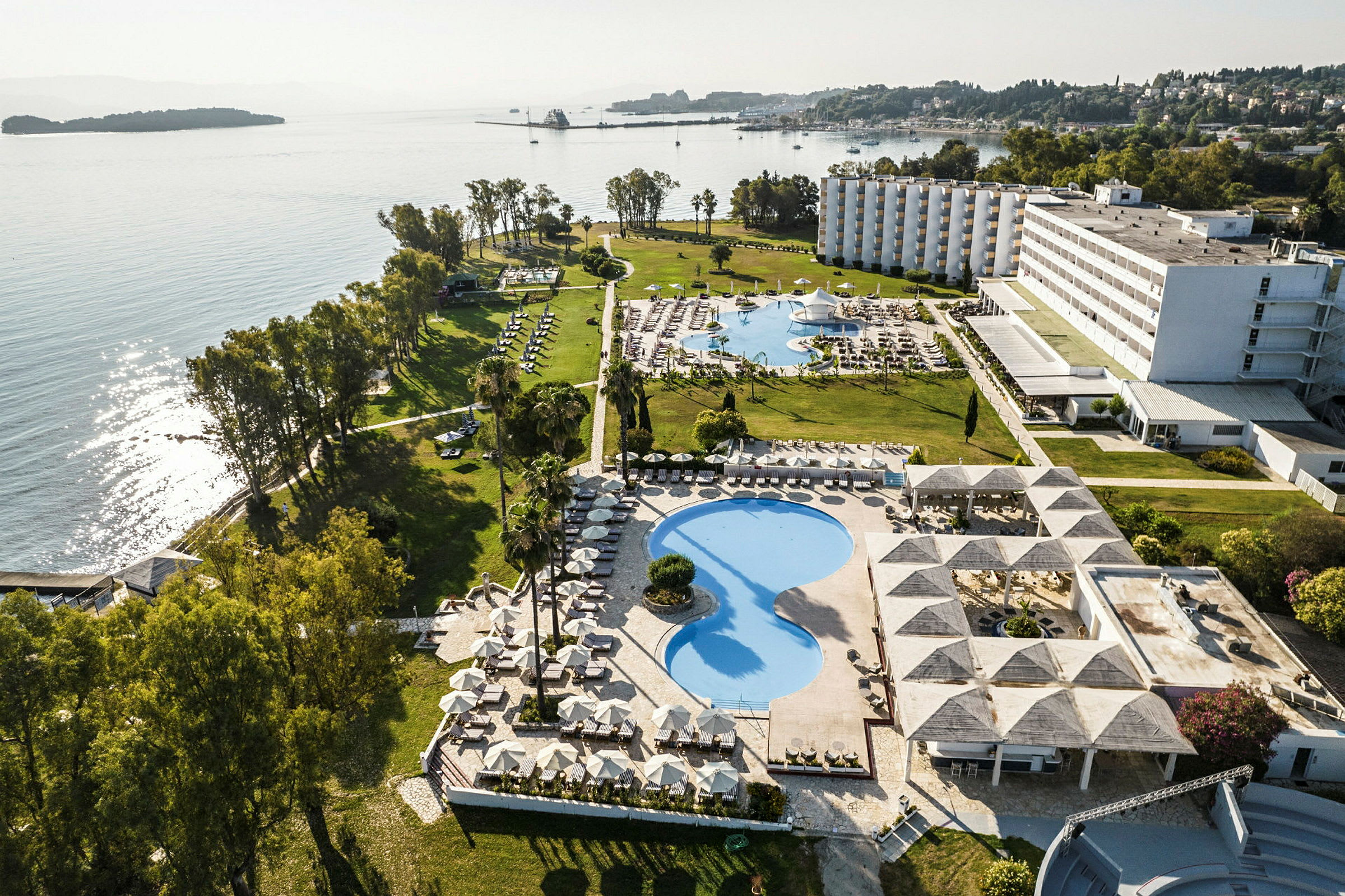Flygvy över ett hotellområde vid havet med pooler, grönområden och byggnader. (AI-genererad text)