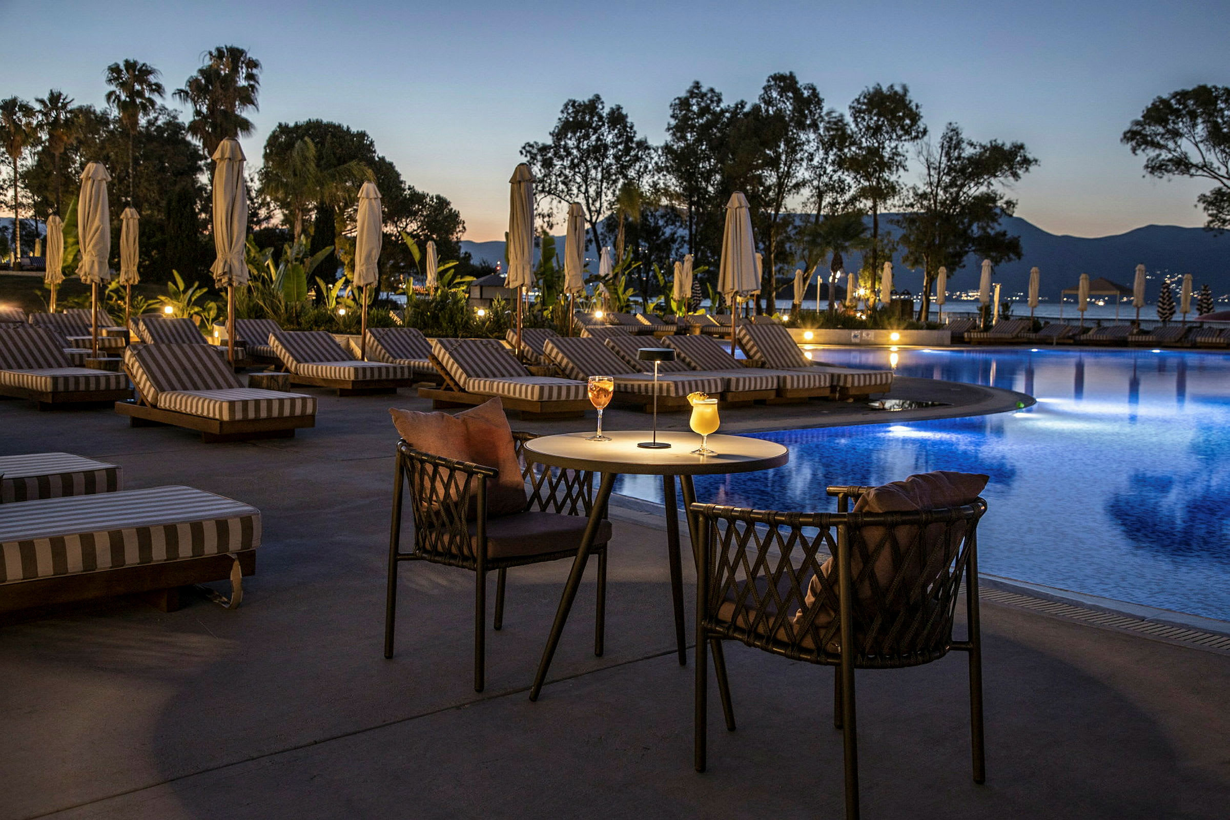 Kvällsvy över en pool med solstolar och ett bord med drycker. (AI-genererad text)