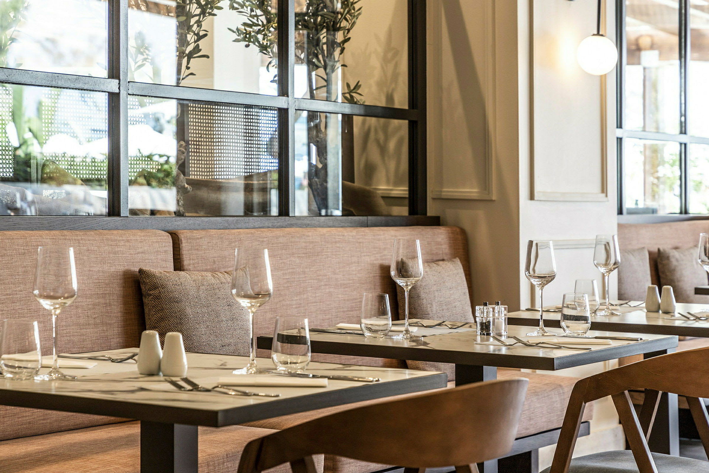 Ett restaurangbord med glas och bestick på en duk, omgiven av stolar och en soffa med kuddar. (AI-genererad text)