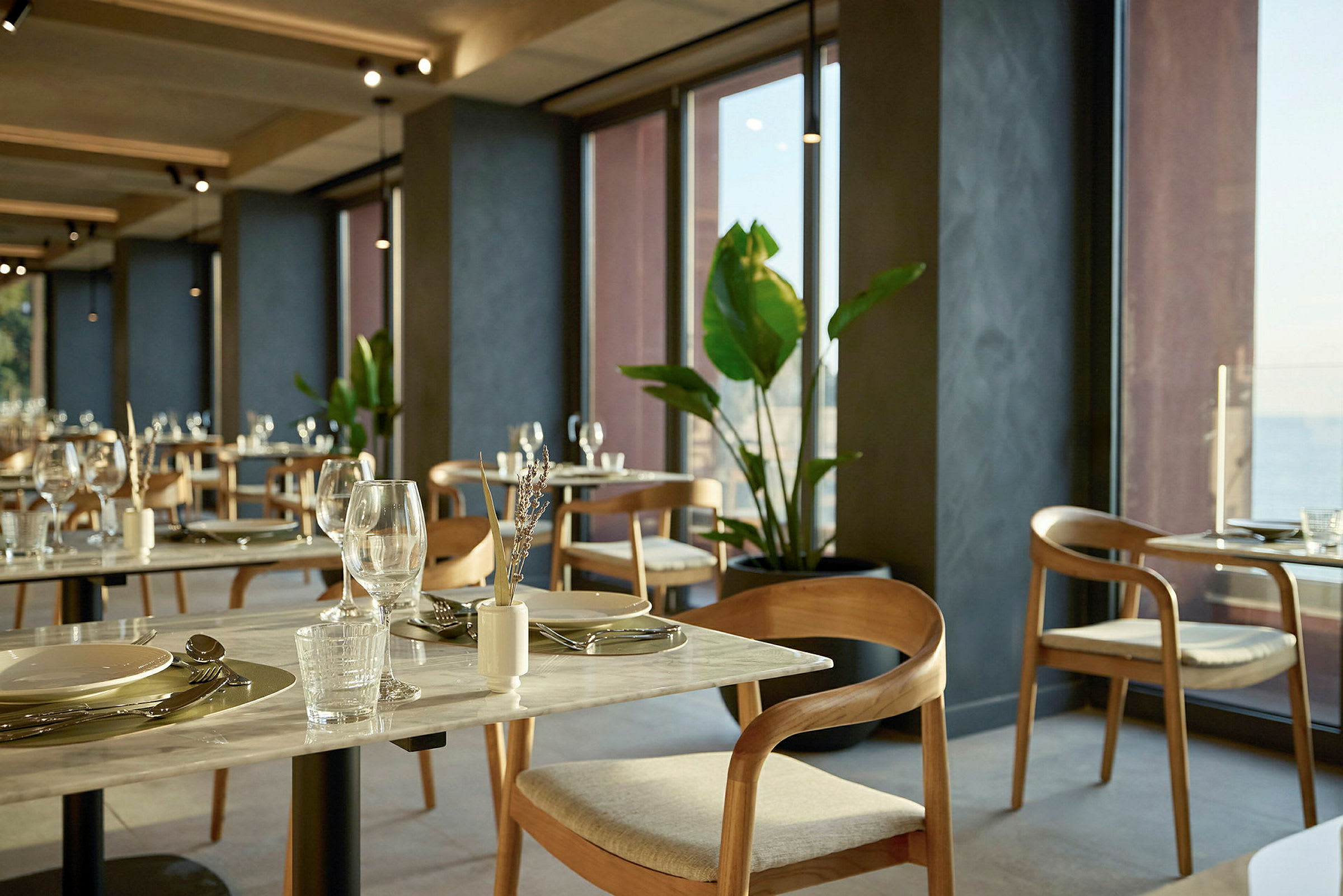 En restaurang med bord och stolar vid stora fönster, dekorerad med växter. (AI-genererad text)