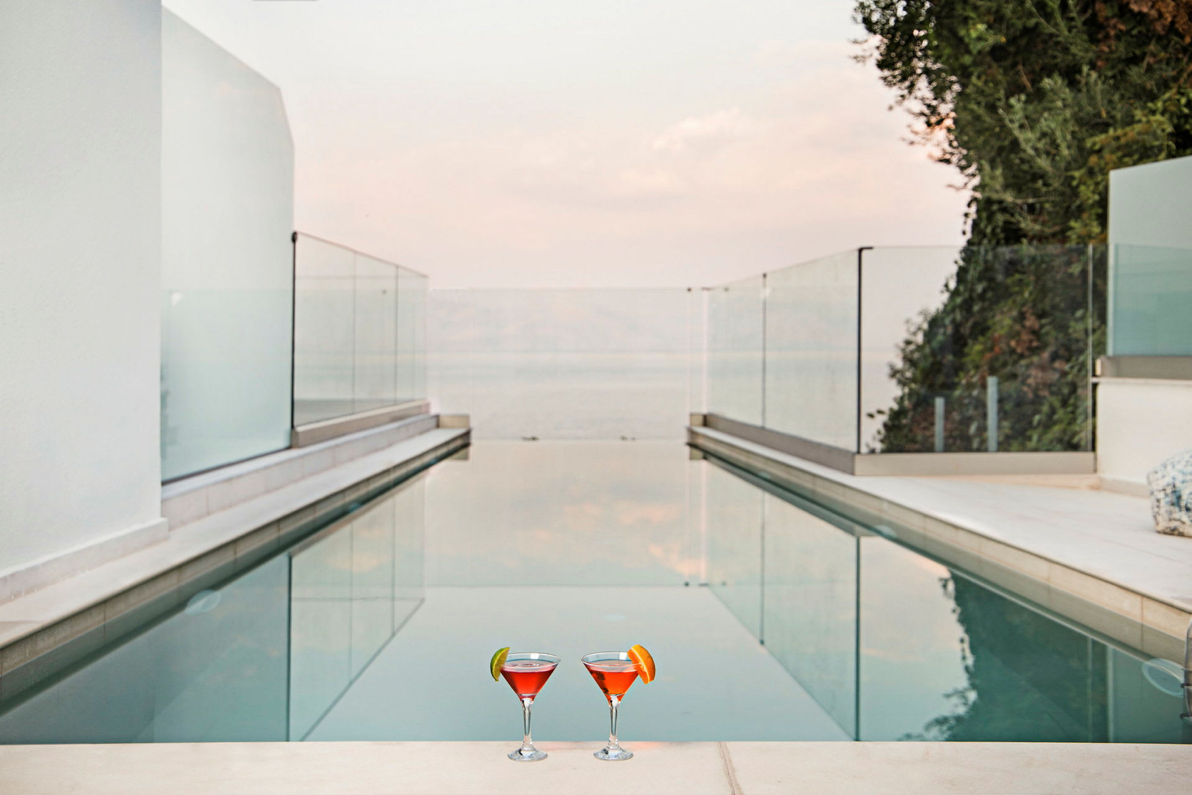 Två glas med orange-röd dryck står vid kanten av en pool omgiven av glasräcken och utsikt över havet. (AI-genererad text)