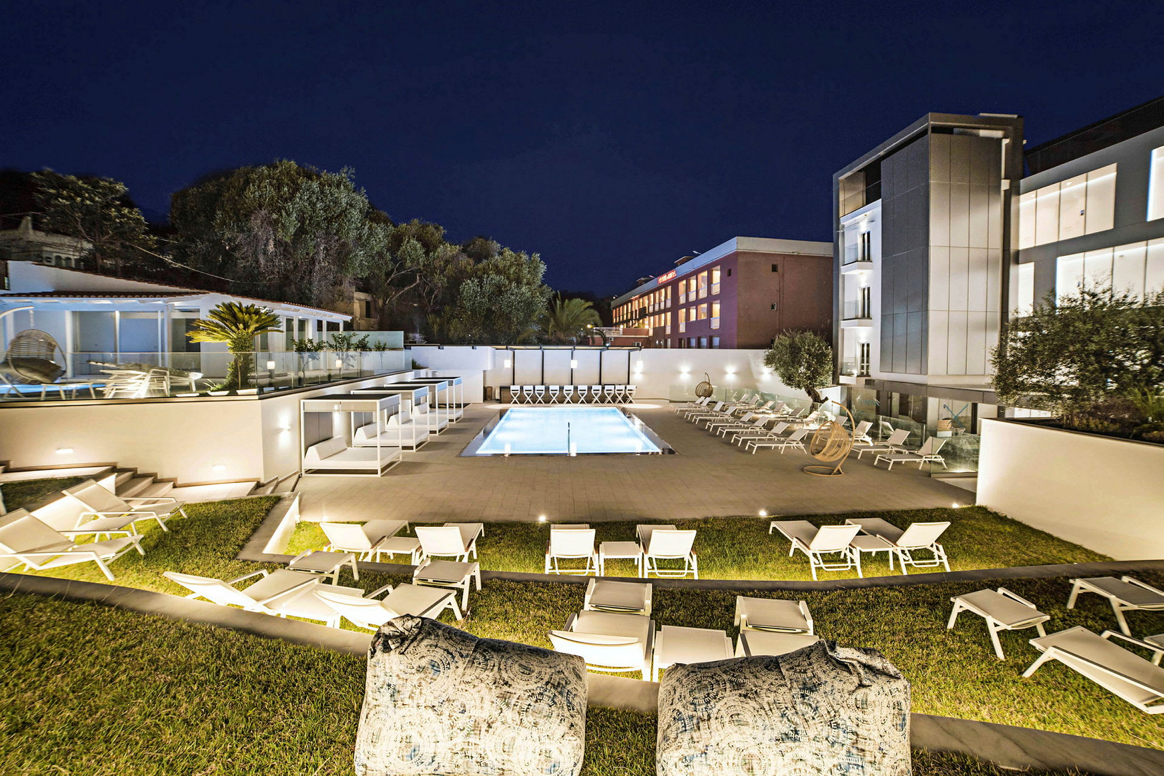 Ett upplyst hotellområde på kvällen, med en pool omgiven av solstolar och moderna byggnader i bakgrunden. (AI-genererad text)