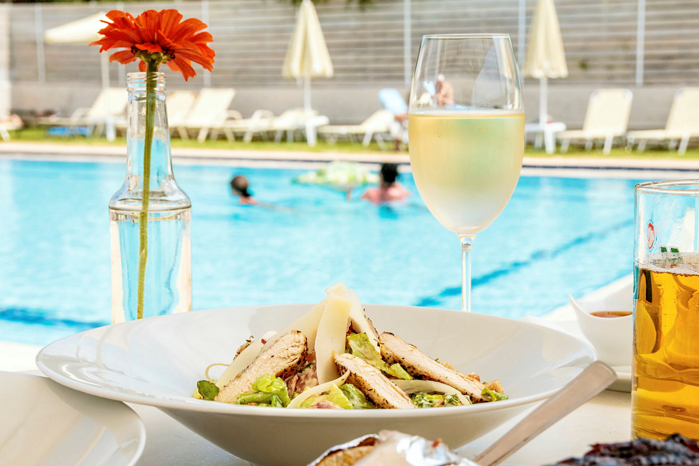 En tallrik med sallad och en glas vitt vin vid en pool. I bakgrunden syns solstolar och personer som badar. (AI-genererad text)