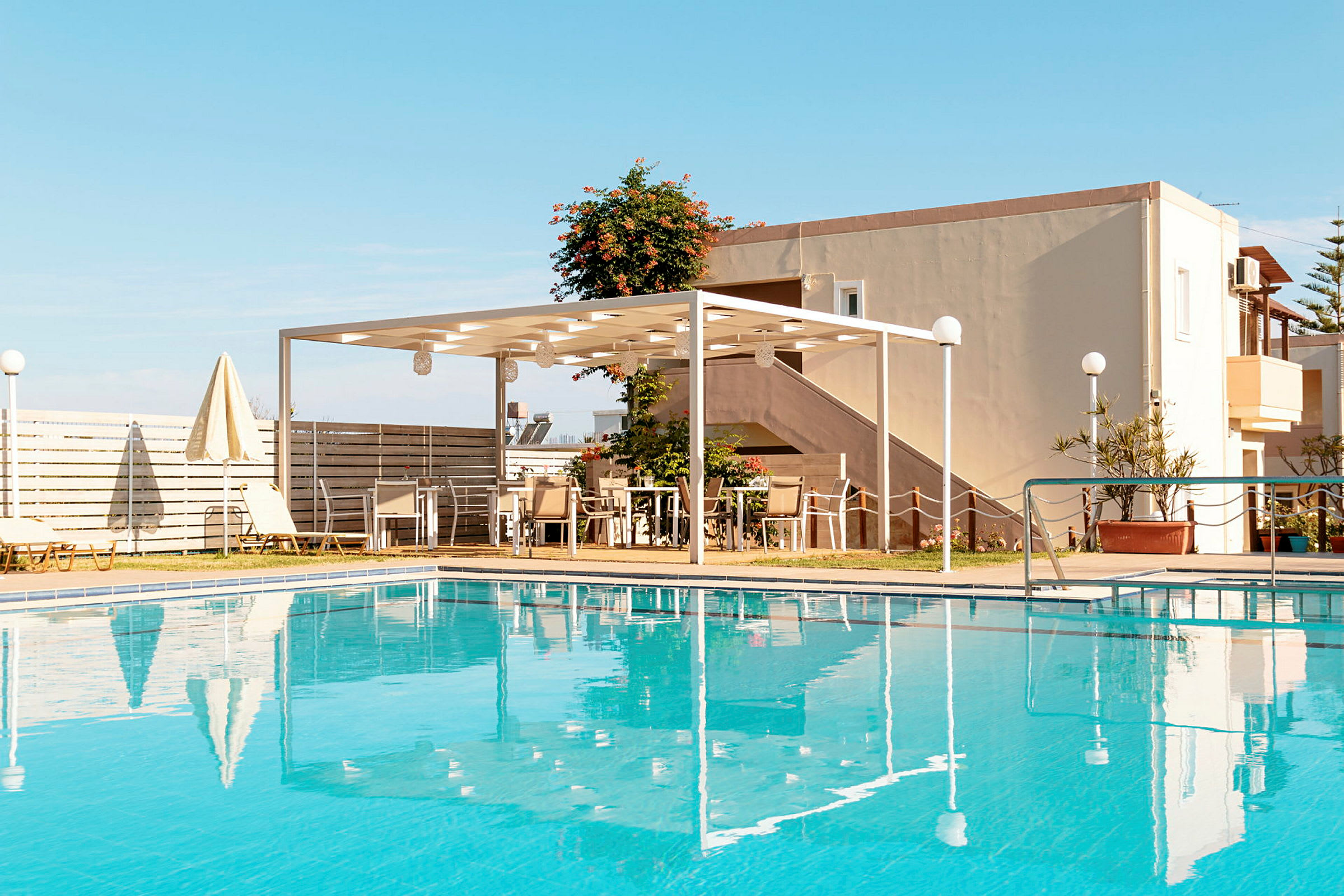 En utomhuspool med klart blått vatten. I bakgrunden syns ett bostadshus, en pergola och några sittplatser under den. (AI-genererad text)
