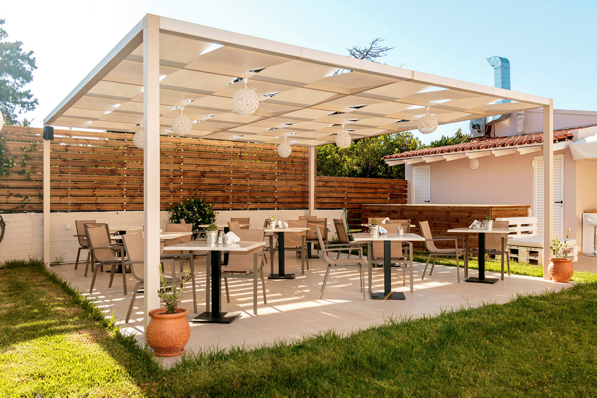 En uteservering med en träpergola som tak, med bord och stolar på ett kaklat golv, omgiven av ett gräsområde och ett staket. (AI-genererad text)