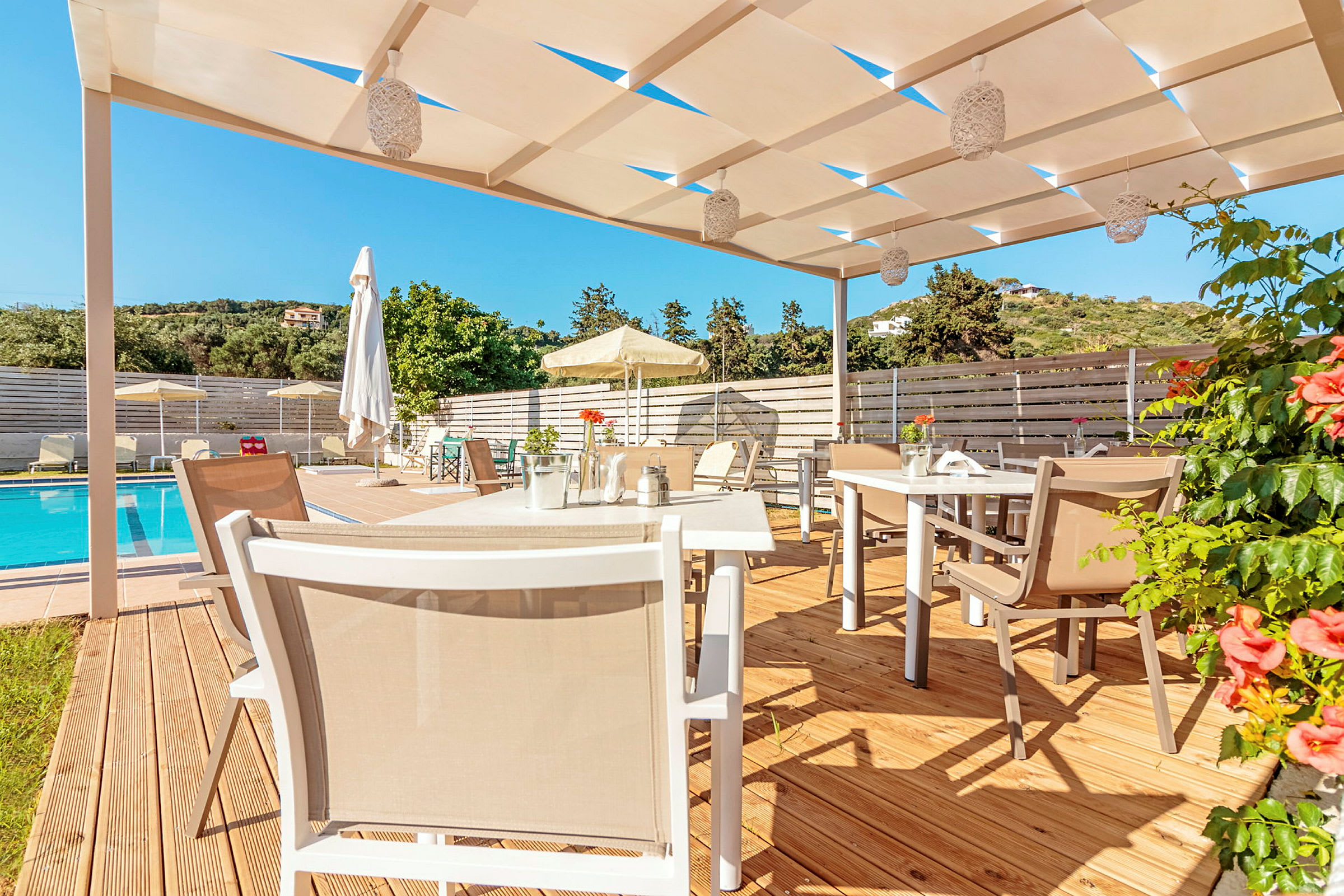 En utomhusterass med bord och stolar under en pergola, nära en pool och omgiven av grönska. (AI-genererad text)