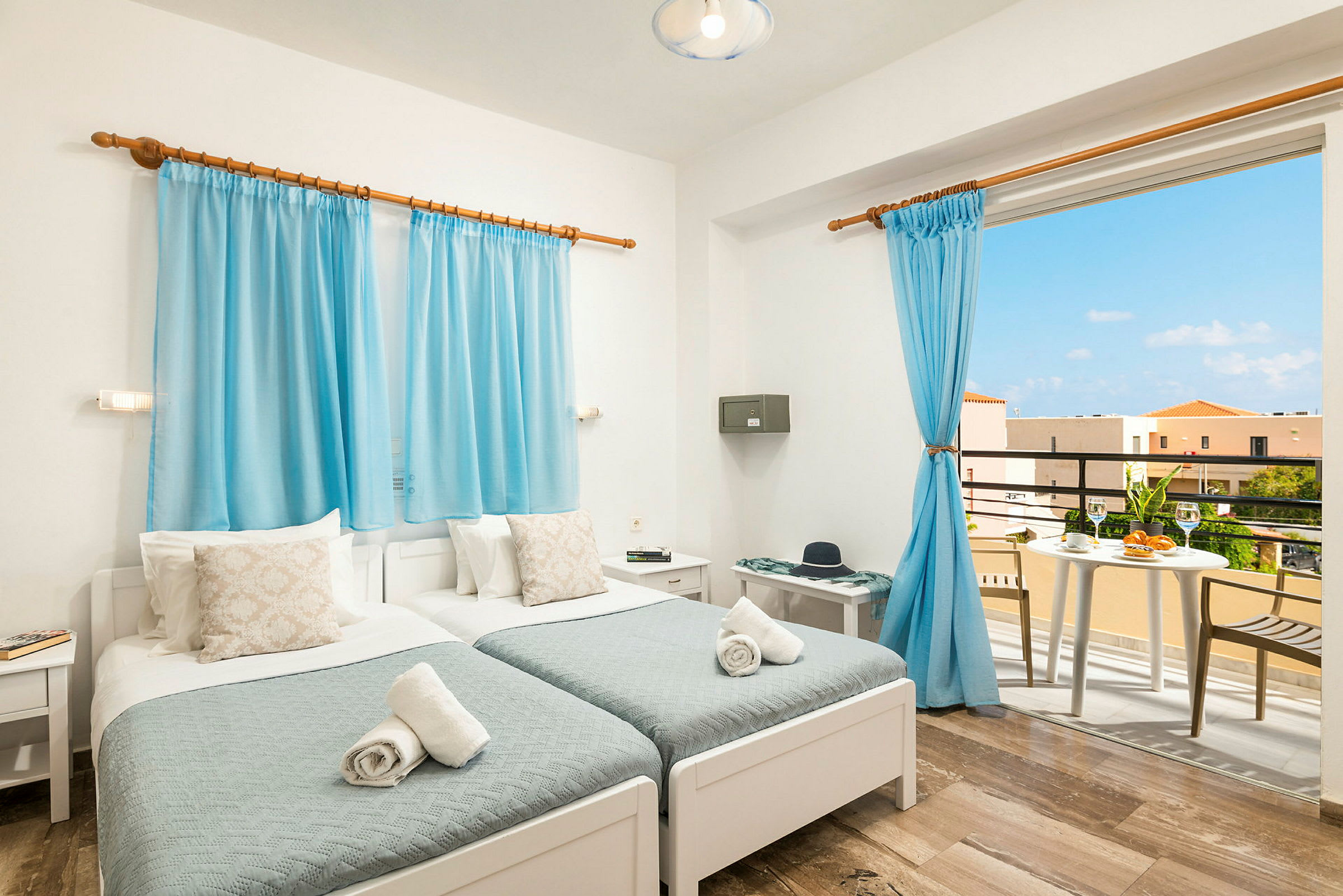Ett ljust och luftigt hotellrum med två enkelsängar, blå gardiner och ett bord på balkongen utanför. (AI-genererad text)