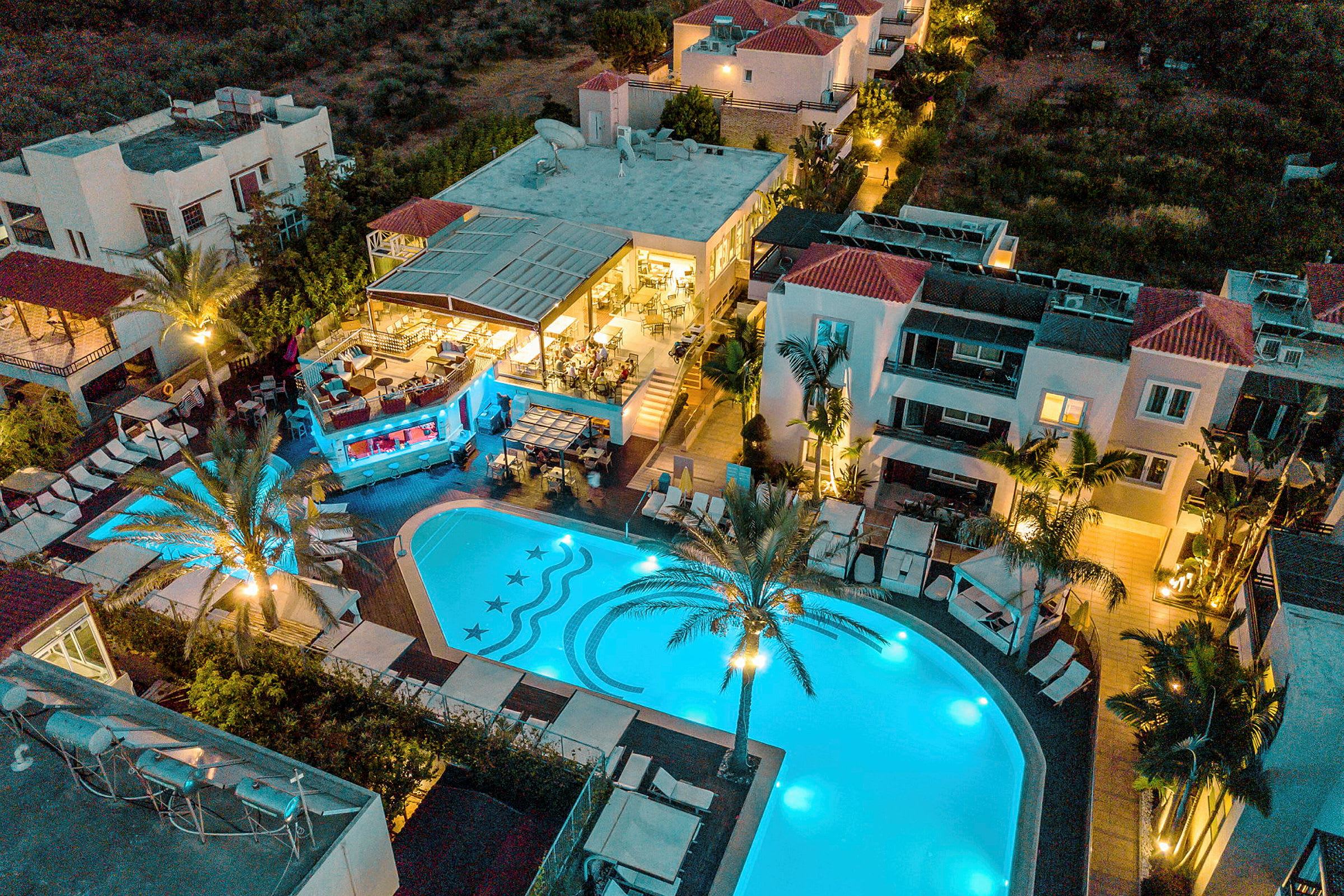 Flygfoto av ett hotellområde med upplyst pool och byggnader runt omkring, en palmliknande växt står bredvid poolen. (AI-genererad text)
