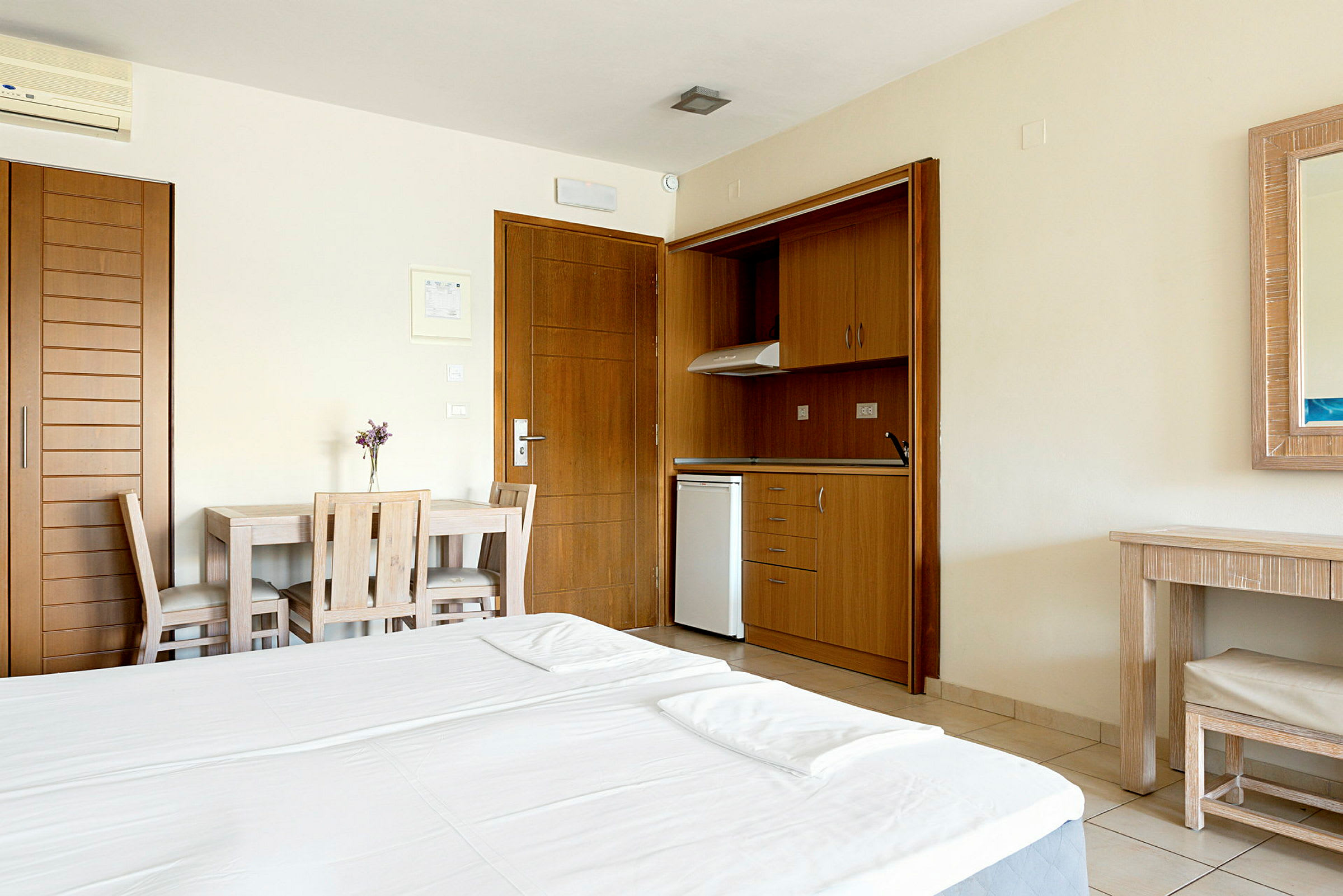 Ett hotellrum med en dubbelsäng, ett litet kök med träinredning, ett matbord med två stolar och en spegel ovanför ett skrivbord. (AI-genererad text)