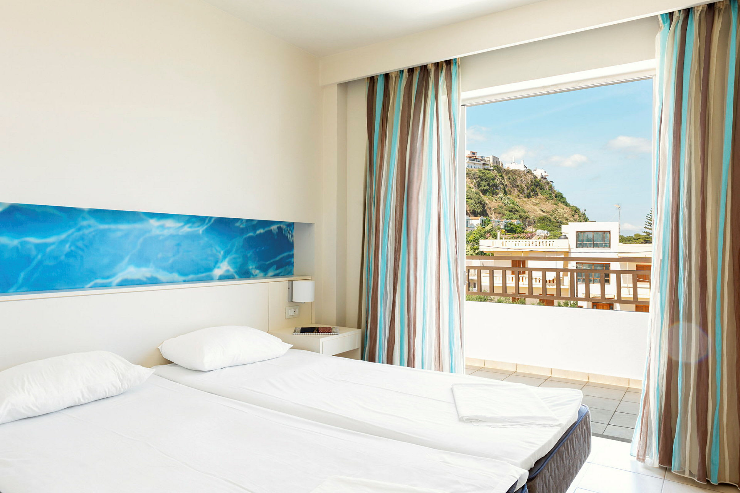 Ett hotellrum med två enkelsängar, ljusa väggar och en blå dekorativ väggpanel. Det finns en stor fönsteröppning med utsikt över en kulle och byggnader utanför. (AI-genererad text)